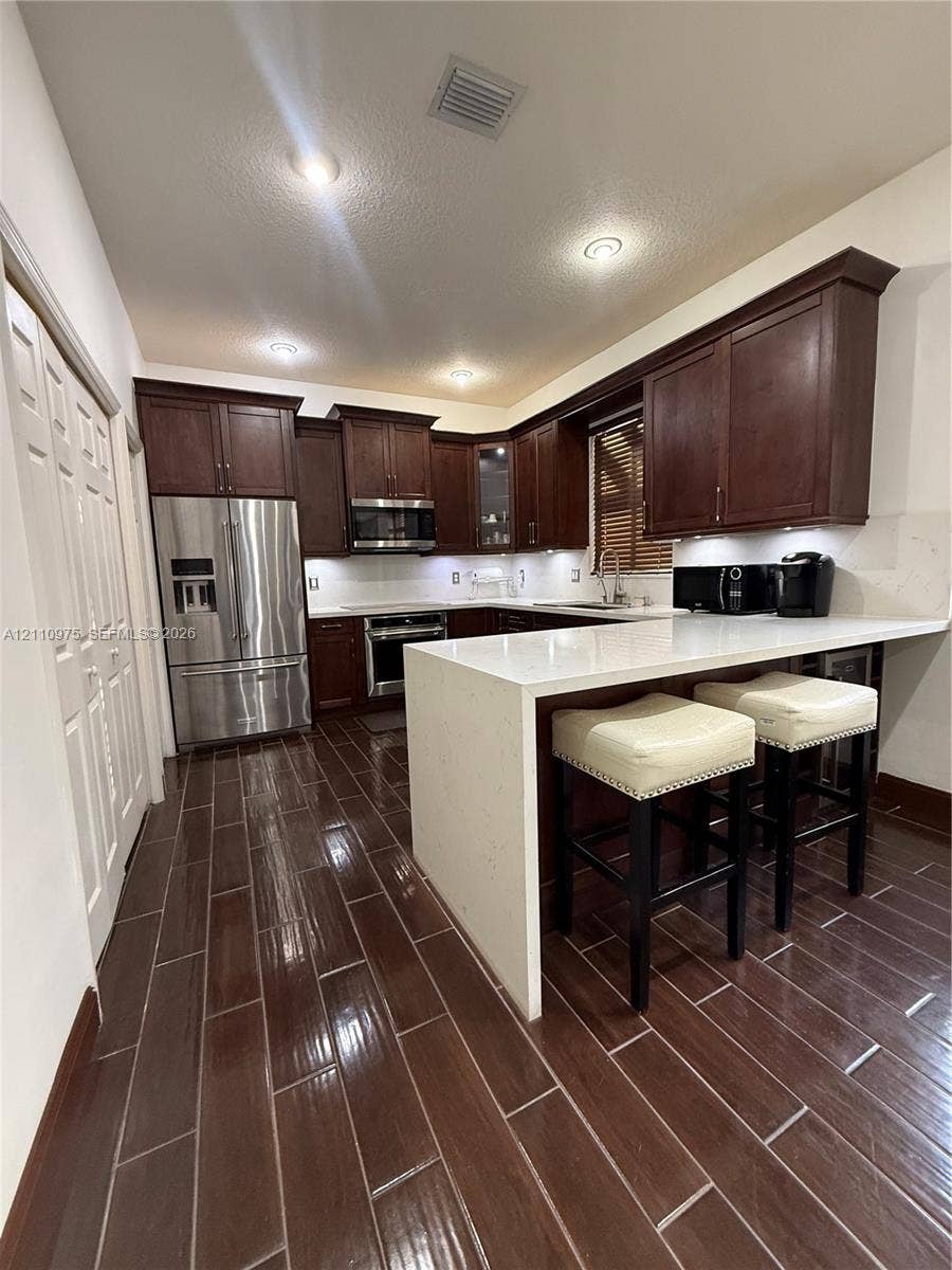 2030 NW 99th Terrace, Pembroke Pines, FL 33024 Kitchen featuring stained wood cabinetry, quartz countertops with a waterfall edge, stainless steel appliances, and dark wood-finish tile flooring