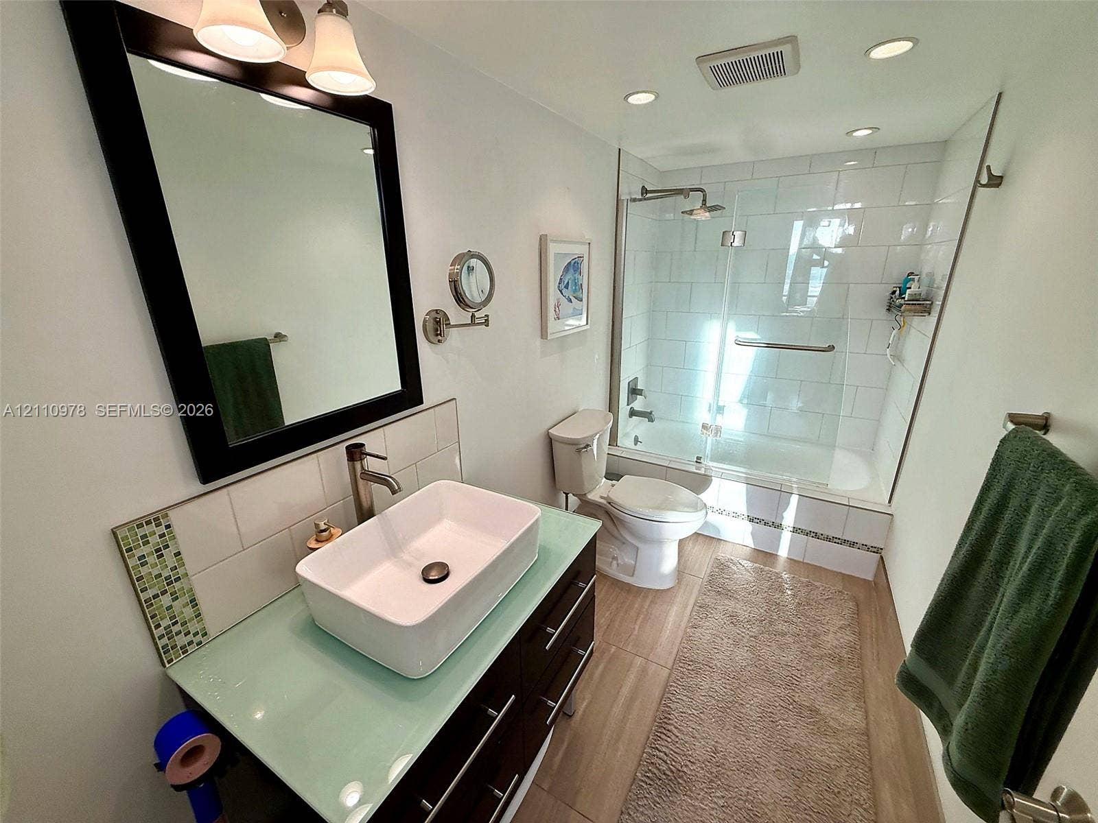 Bathroom featuring a dark wood-finish vanity with a glass countertop, rectangular vessel sink, white subway tile shower surround, and wood-finish flooring