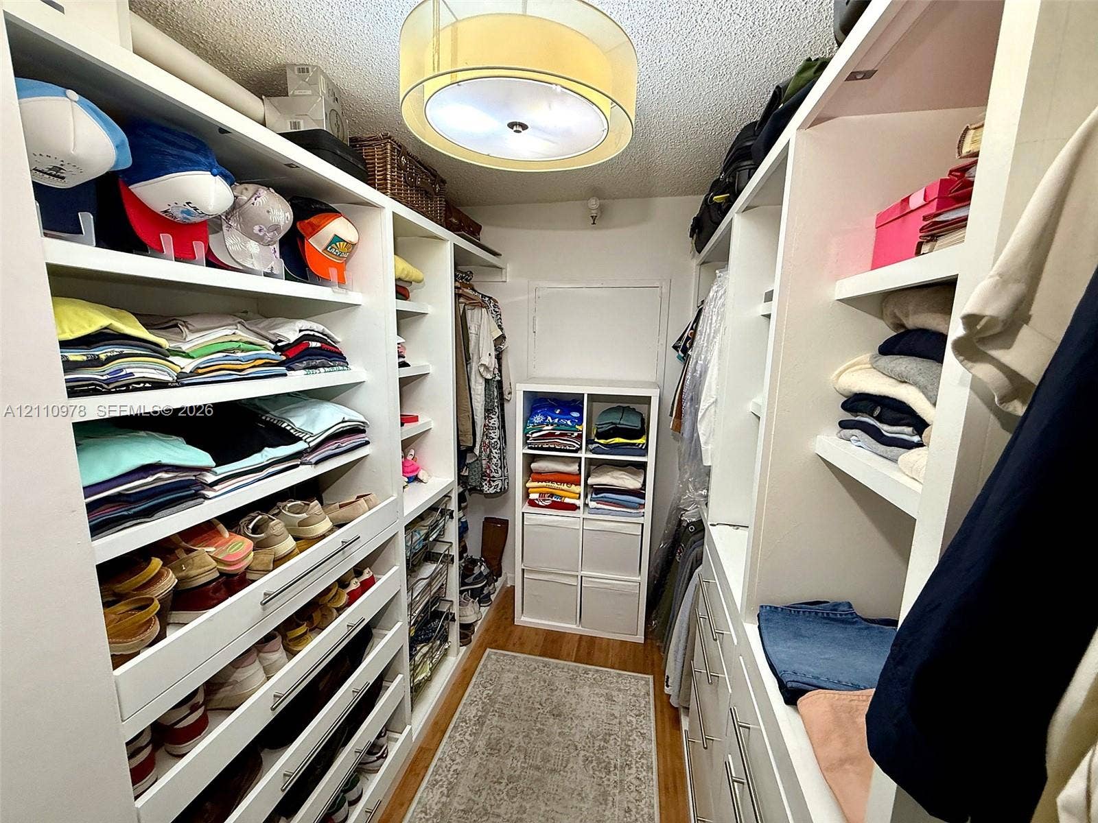Walk-in closet featuring white built-in shelving, integrated dresser drawers with silver-tone hardware, wood-finish flooring, and a drum-style ceiling fixture
