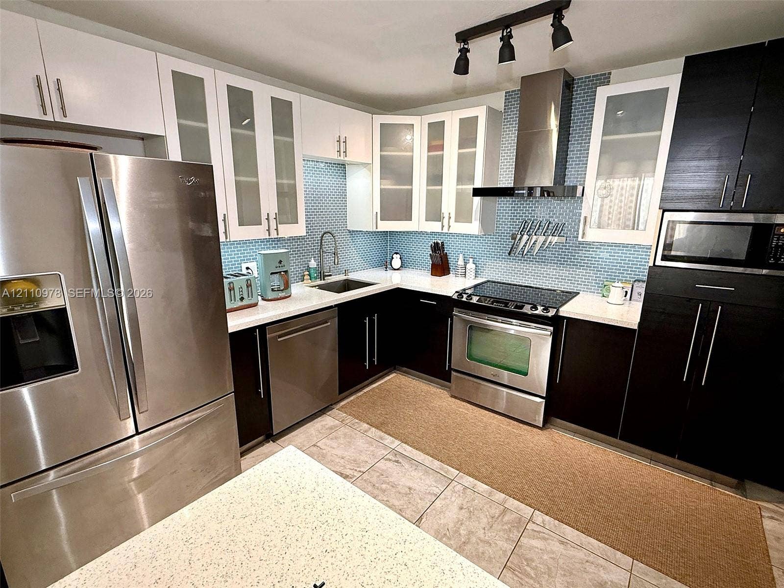 Kitchen featuring two-tone cabinetry, stainless steel appliances, blue glass-tiled backsplash, light-colored stone-finish countertops, and overhead track lighting