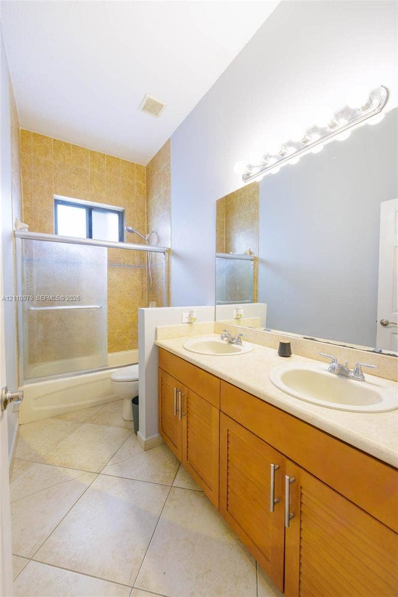 Dual-sink vanity with light-tone countertop, wood-finish cabinetry, full-width mirror, and tiled shower enclosure with sliding glass door