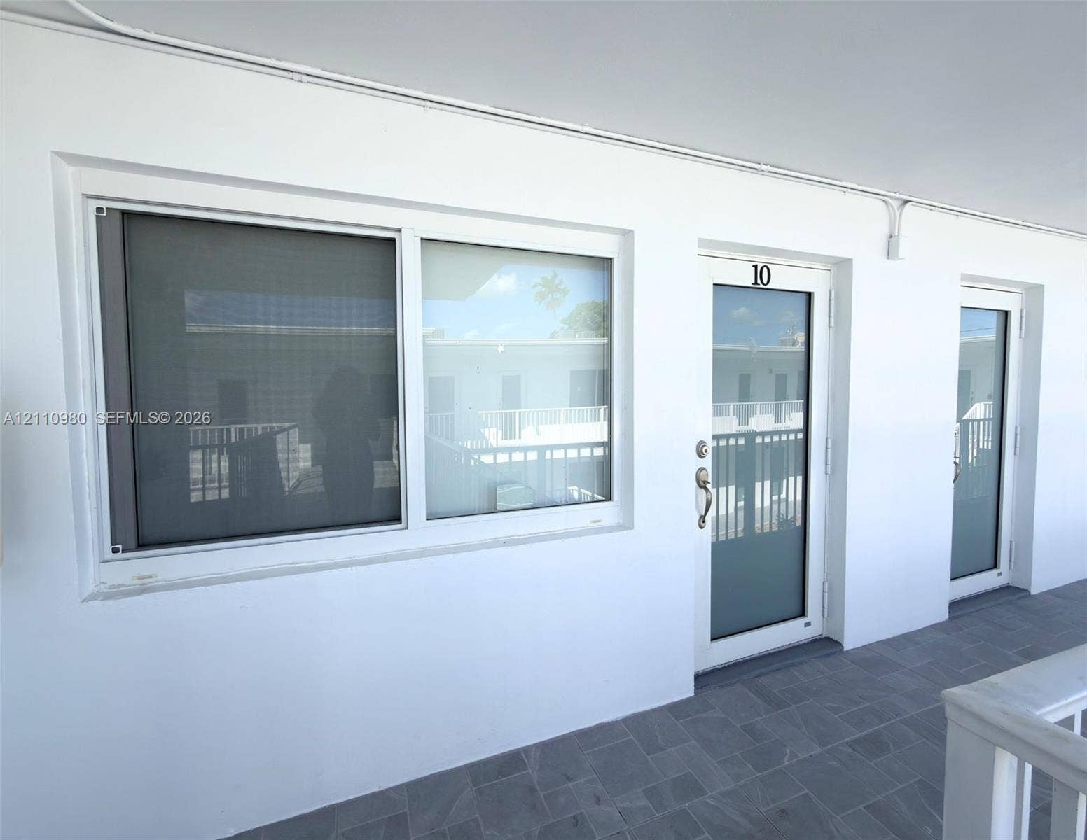 Exterior wall featuring white stucco finish, sliding glass window with metal screen, and white-framed glass entry doors with brushed metal hardware, complemented by gray tile flooring