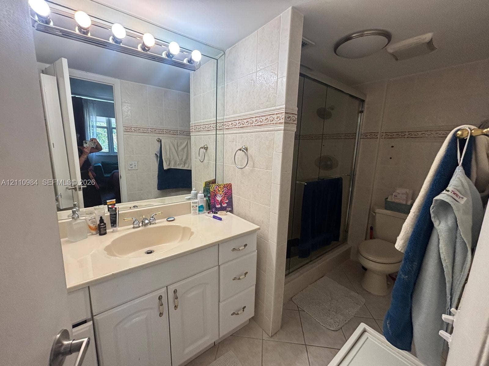 3745 NE 171st Street, #77, North Miami Beach, North Miami Beach, FL 33160 Bathroom featuring a white vanity cabinet with an integrated sink, a large wall-mounted mirror with an overhead five-light fixture, ceramic wall tiling with a decorative border, and a glass-enclosed shower