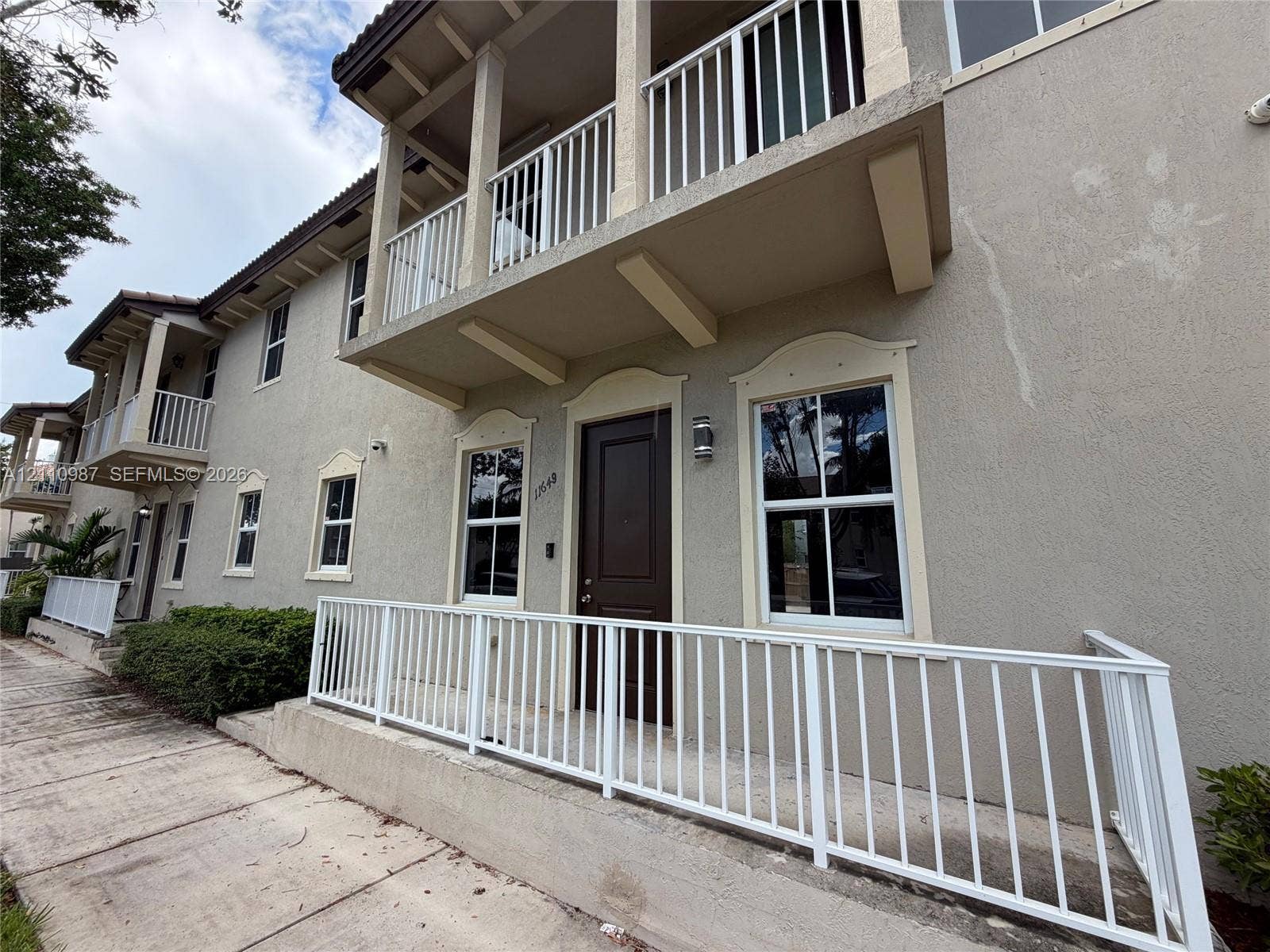 Stucco exterior featuring a second-story balcony with white railing, covered ground-level porch, decorative window trim, and a concrete walkway