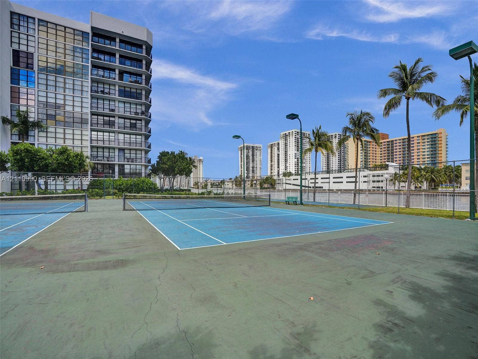 800 Parkview Drive, #131, Hallandale Beach, FL 33009 Hard-court tennis facility featuring perimeter chain-link fencing, dual court lighting, and mature palm trees