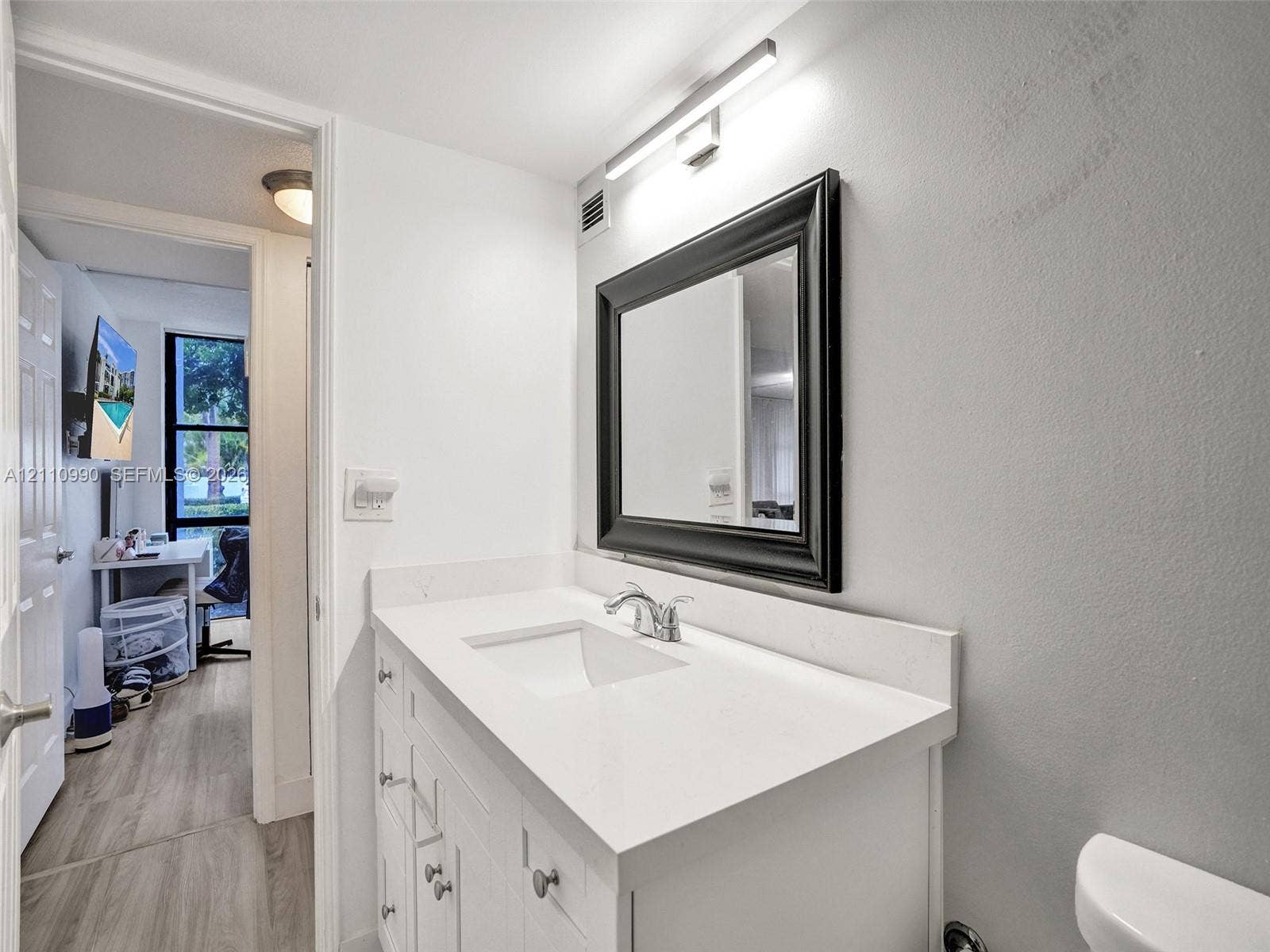 800 Parkview Drive, #131, Hallandale Beach, FL 33009 White vanity with a solid-surface countertop and integrated sink, complemented by a framed mirror, wall-mounted vanity lighting, and matte-finish hardware