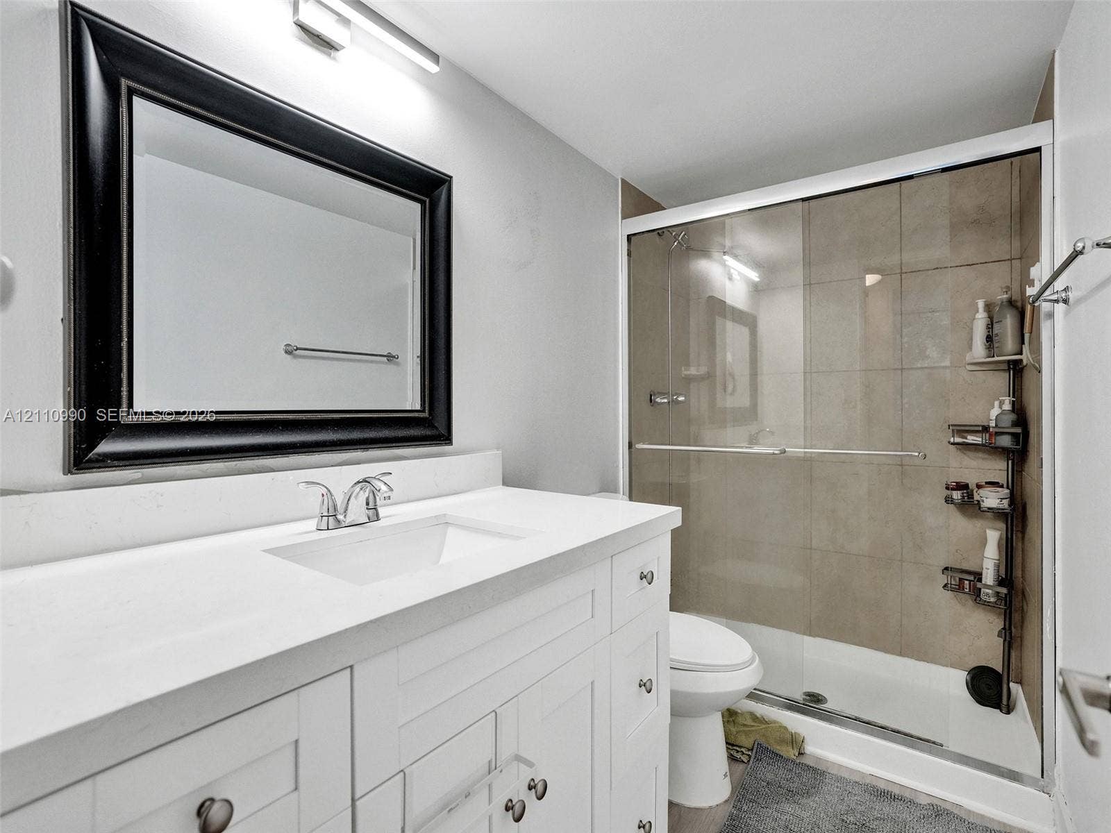 800 Parkview Drive, #131, Hallandale Beach, FL 33009 White shaker-style vanity with a solid surface countertop, rectangular integrated sink, chrome faucet, framed mirror, and tiled shower enclosure with a glass sliding door