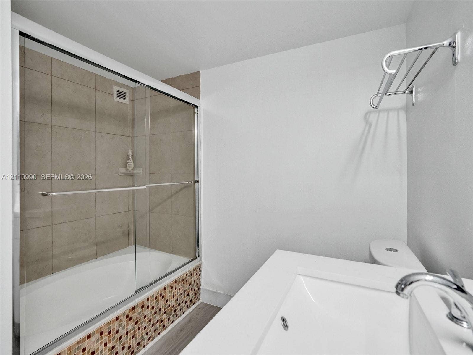 800 Parkview Drive, #131, Hallandale Beach, FL 33009 Bathtub with a glass sliding enclosure, tan tile surround, multicolored mosaic tile apron, white vanity with an integrated basin, and chrome wall-mounted towel rack