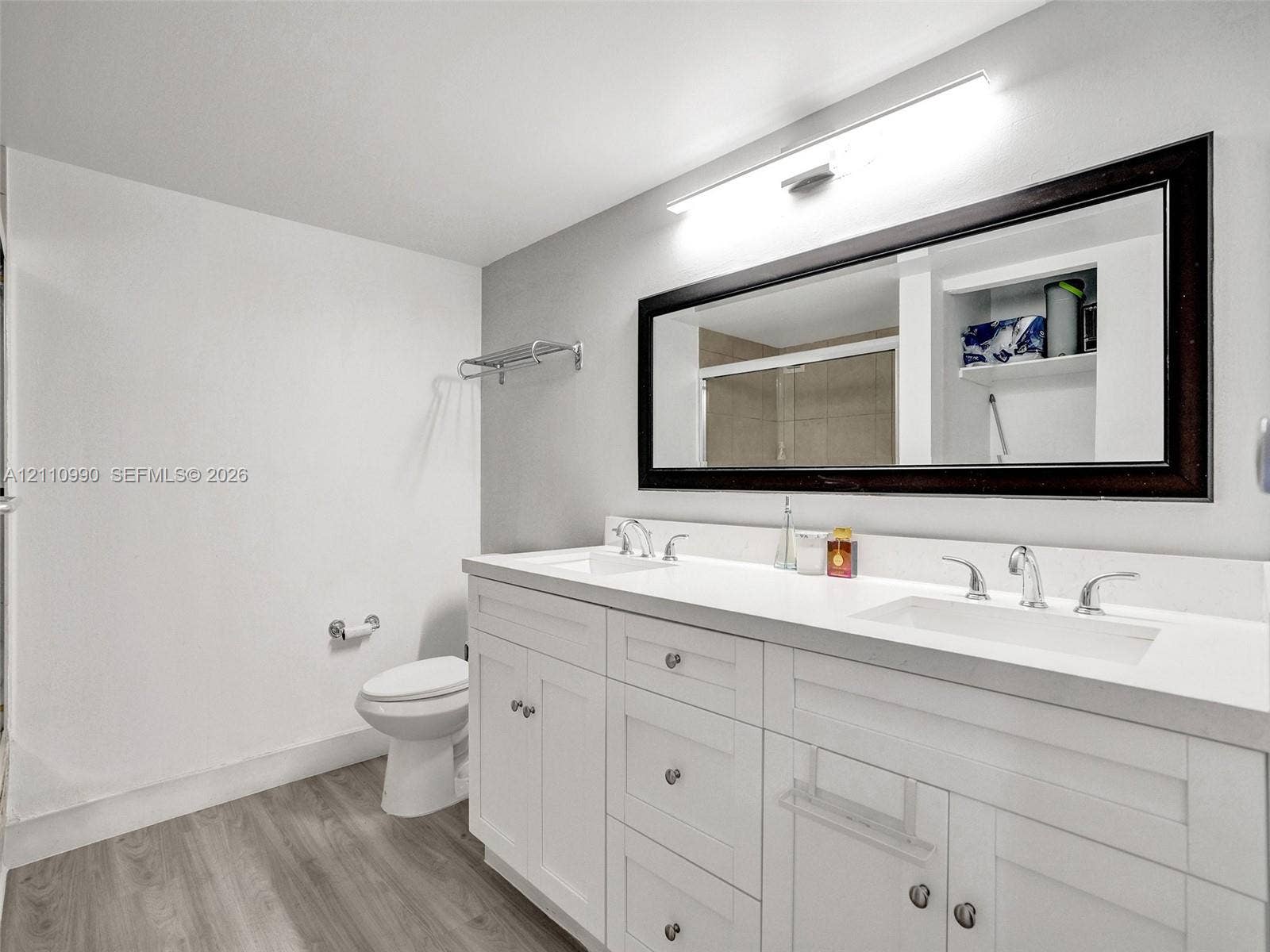 800 Parkview Drive, #131, Hallandale Beach, FL 33009 White dual-sink vanity with a solid surface countertop, wood-finish flooring, full-width mirror with a dark frame, and chrome-finish hardware