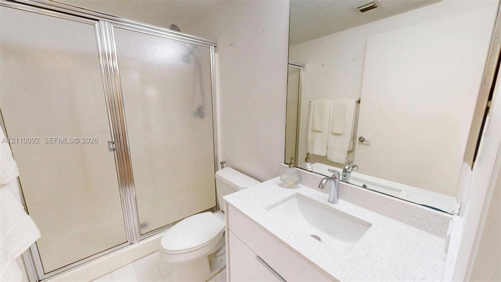 Bathroom featuring a vanity with a white composite countertop, integrated sink, chrome faucet, full-width mirror, and a glass-enclosed shower with a chrome frame