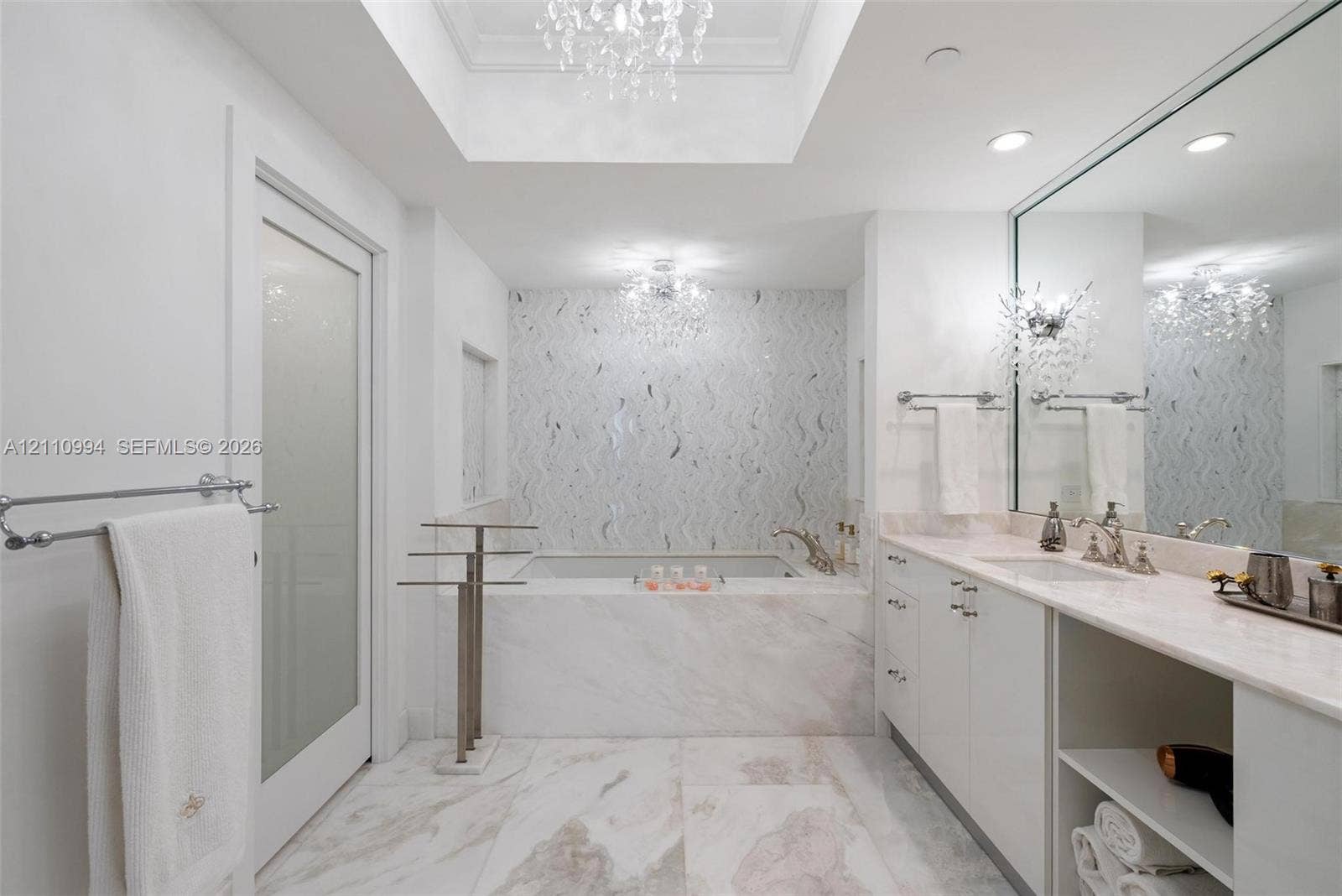 17875 Collins Avenue, #2405, Sunny Isles Beach, Sunny Isles Beach, FL 33160 Soaking tub with stone surround, custom tiled accent wall, skylight with recessed tray ceiling, dual crystal chandeliers, and white cabinetry with stone countertop