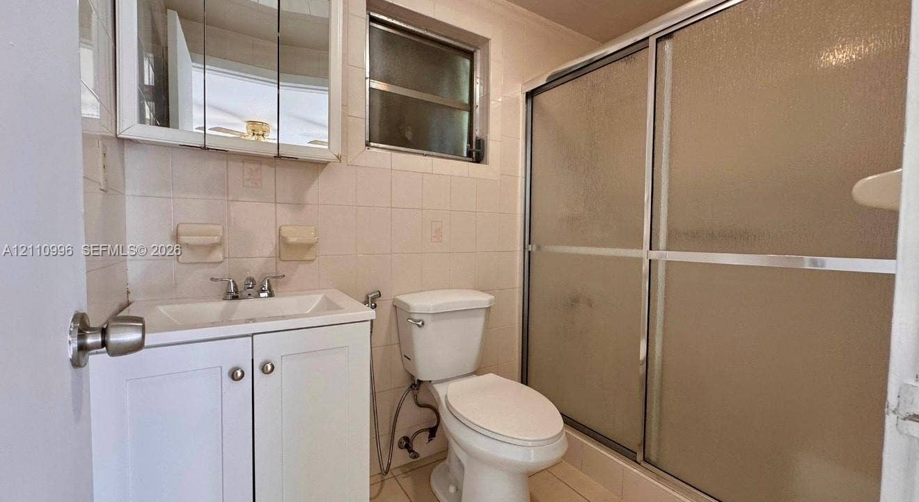Bathroom featuring a white vanity cabinet, a wall-mounted medicine cabinet with a mirror, a ceramic tile shower enclosure with sliding glass doors, and white wall tile