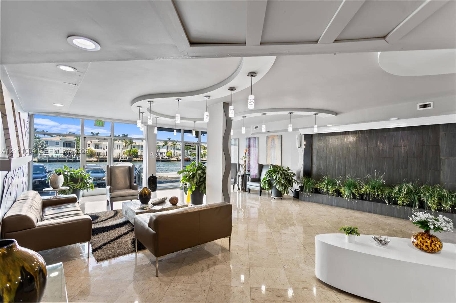 Expansive open-concept space featuring polished stone flooring, a decorative stone-tiled water feature, curvilinear coffered ceiling accents, and floor-to-ceiling glass windows with water frontage