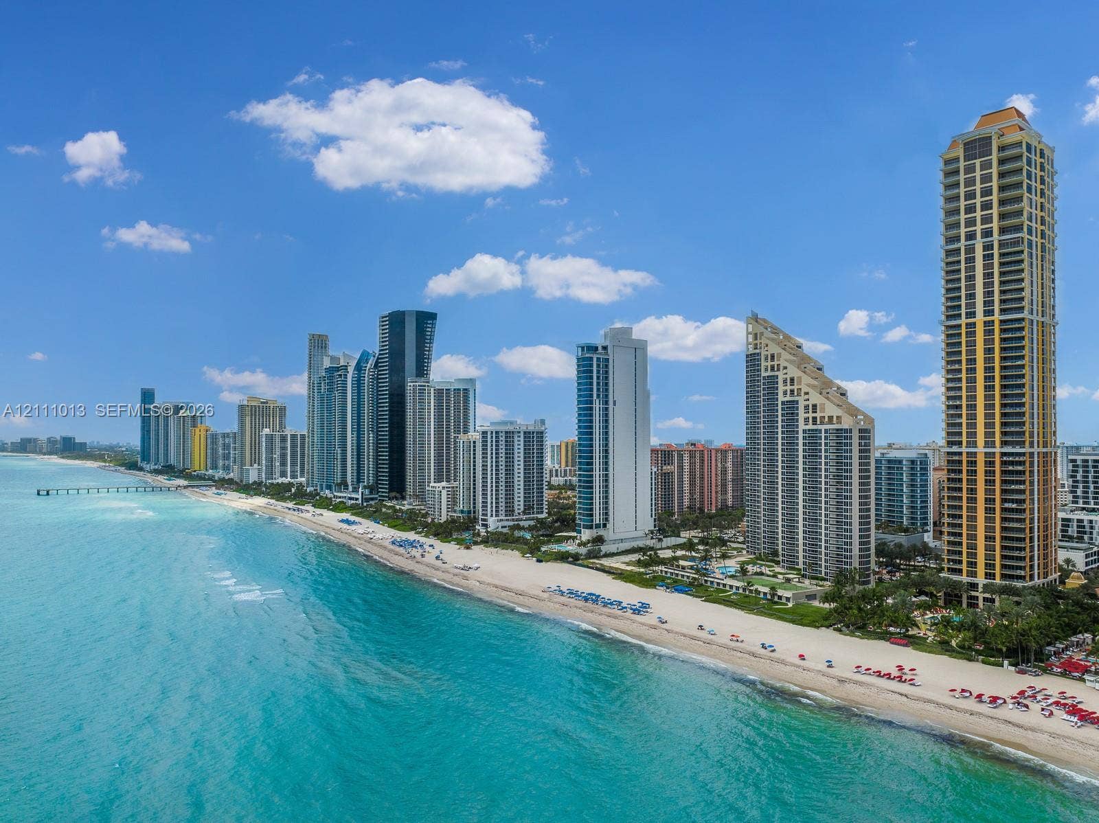 Aerial coastal perspective featuring a stretch of sandy beach, turquoise ocean waters, and high-rise structures