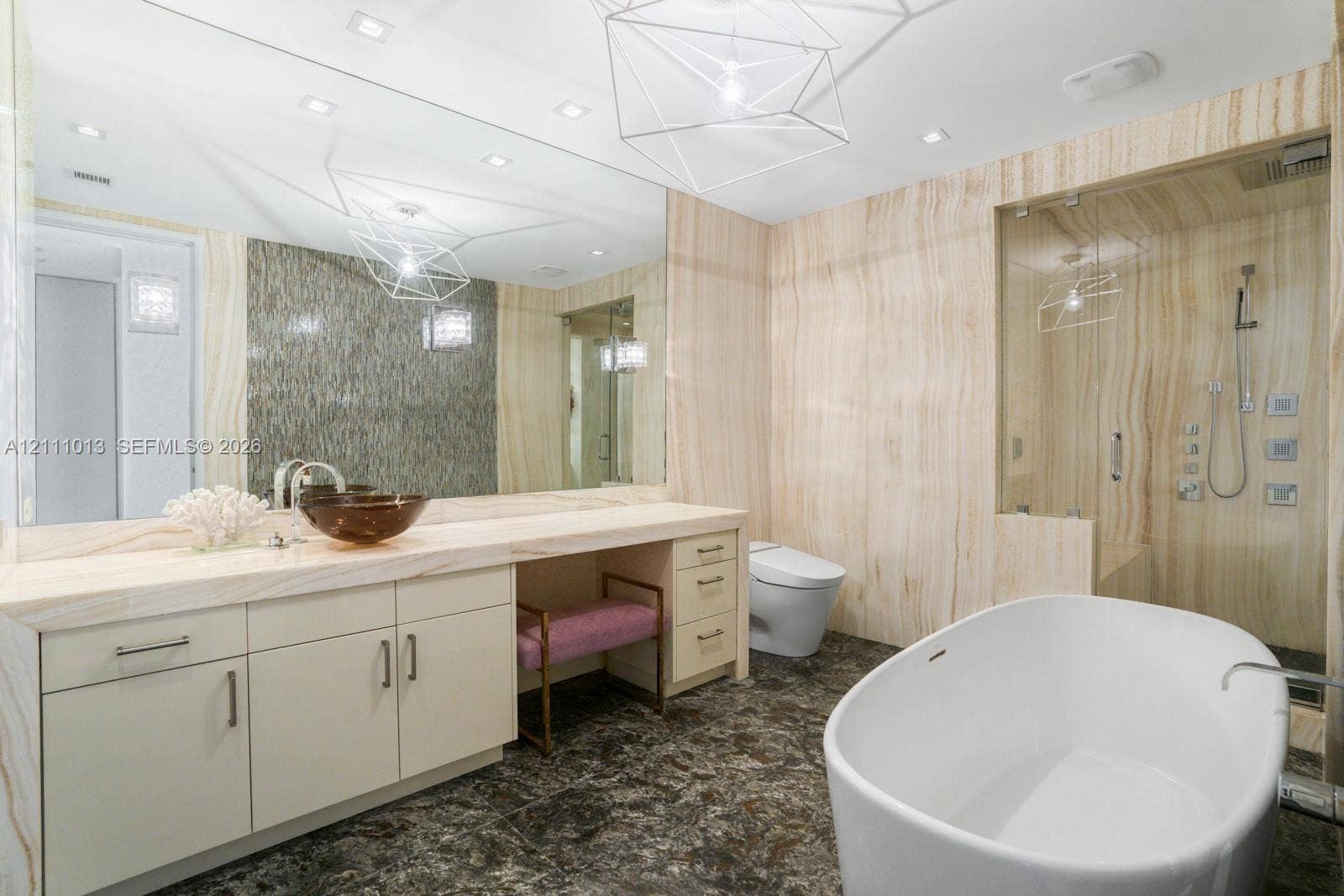 Custom vanity with stone countertop and vessel sink, floor-to-ceiling stone wall cladding, walk-in glass shower with multi-head fixture, freestanding soaking tub, and geometric pendant lighting