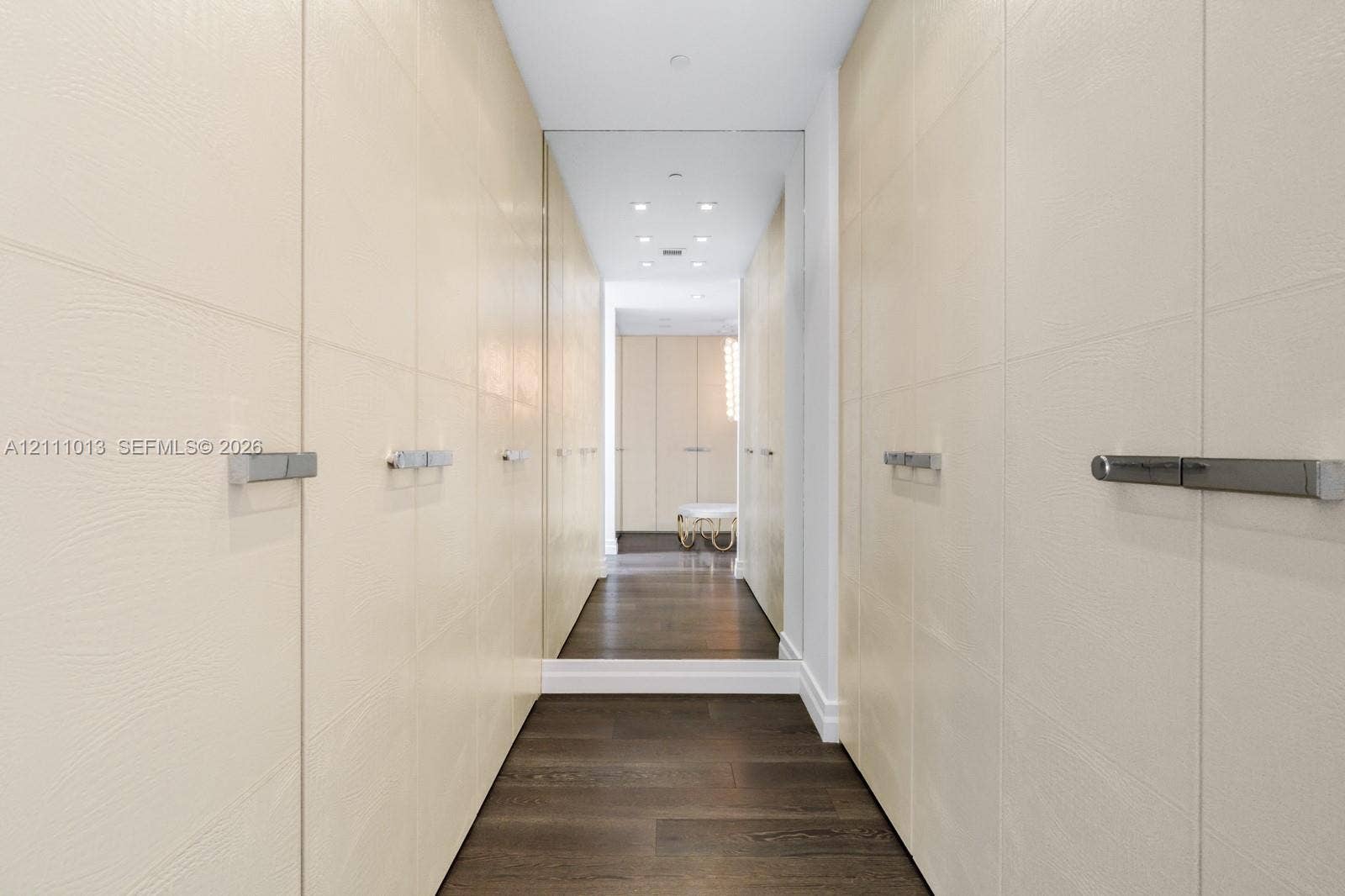 Custom floor-to-ceiling cabinetry with textured cream-colored finish, polished metal hardware, full-height wall mirrors, recessed ceiling lighting, and dark wood-finish flooring