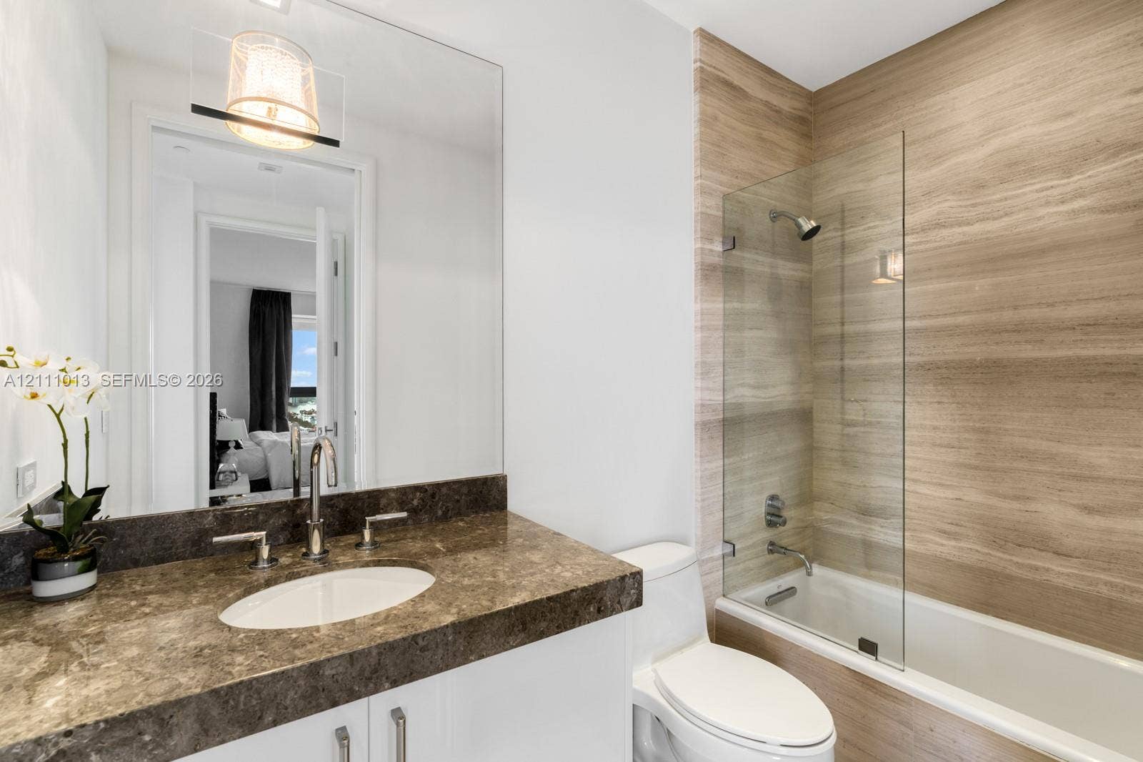 Bathroom featuring a stone-topped vanity with an undermount sink, large wall-mounted mirror, white cabinetry, and a shower-tub combination with a glass panel and stone-finish tile surround