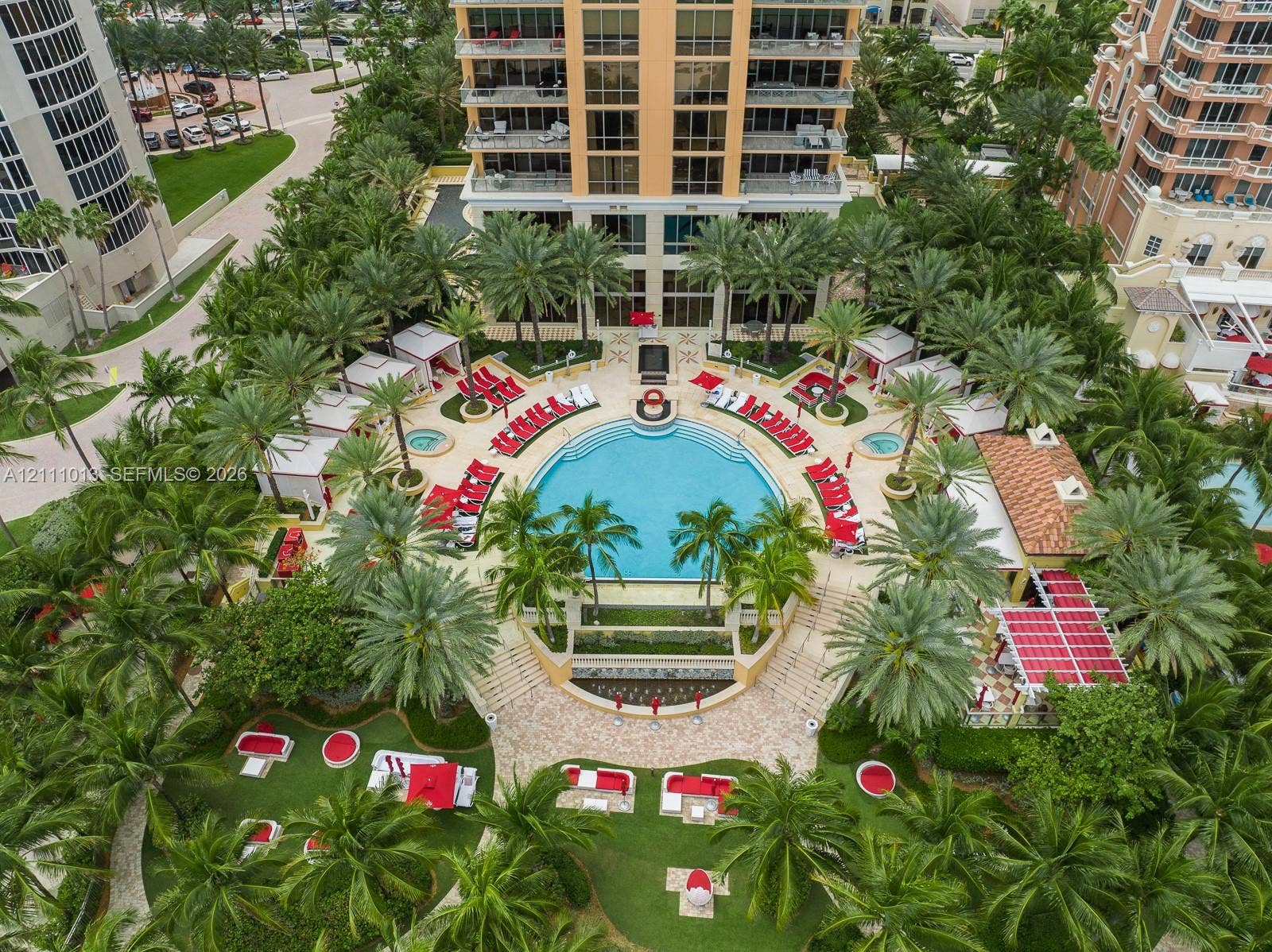 Resort-style pool area featuring a circular swimming pool, surrounding cabanas, manicured landscaping with mature palm trees, and decorative paved walkways