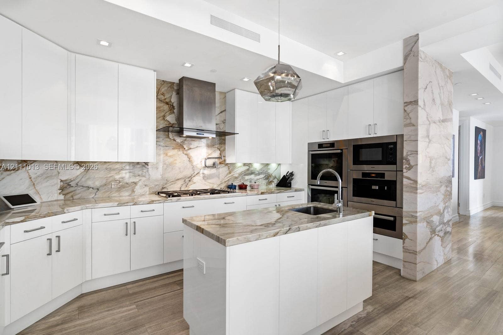 Contemporary kitchen featuring high-gloss white cabinetry, expansive stone slab countertops and backsplash, integrated stainless steel double wall ovens, wood-finish flooring, and a central island with an undermount sink