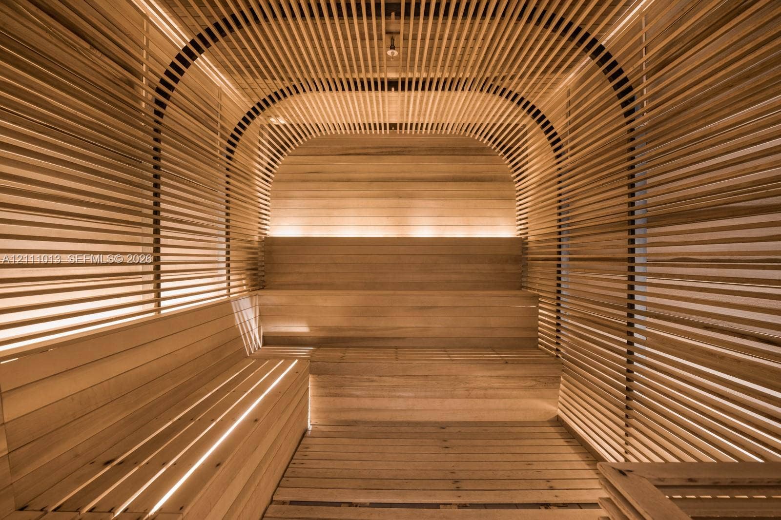 Sauna featuring light-toned wood slat wall paneling, tiered bench seating, integrated strip lighting, and a curved architectural ceiling design