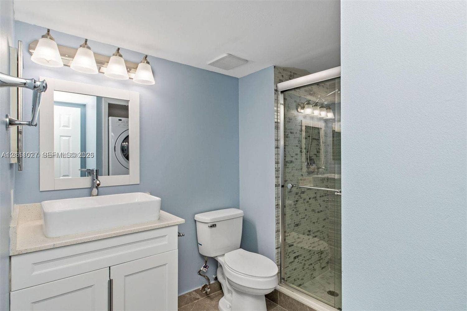 Bathroom featuring a vanity with a white rectangular vessel sink, stone-finish countertop, four-light wall fixture, framed mirror, and a walk-in shower with a glass door and tiled surround