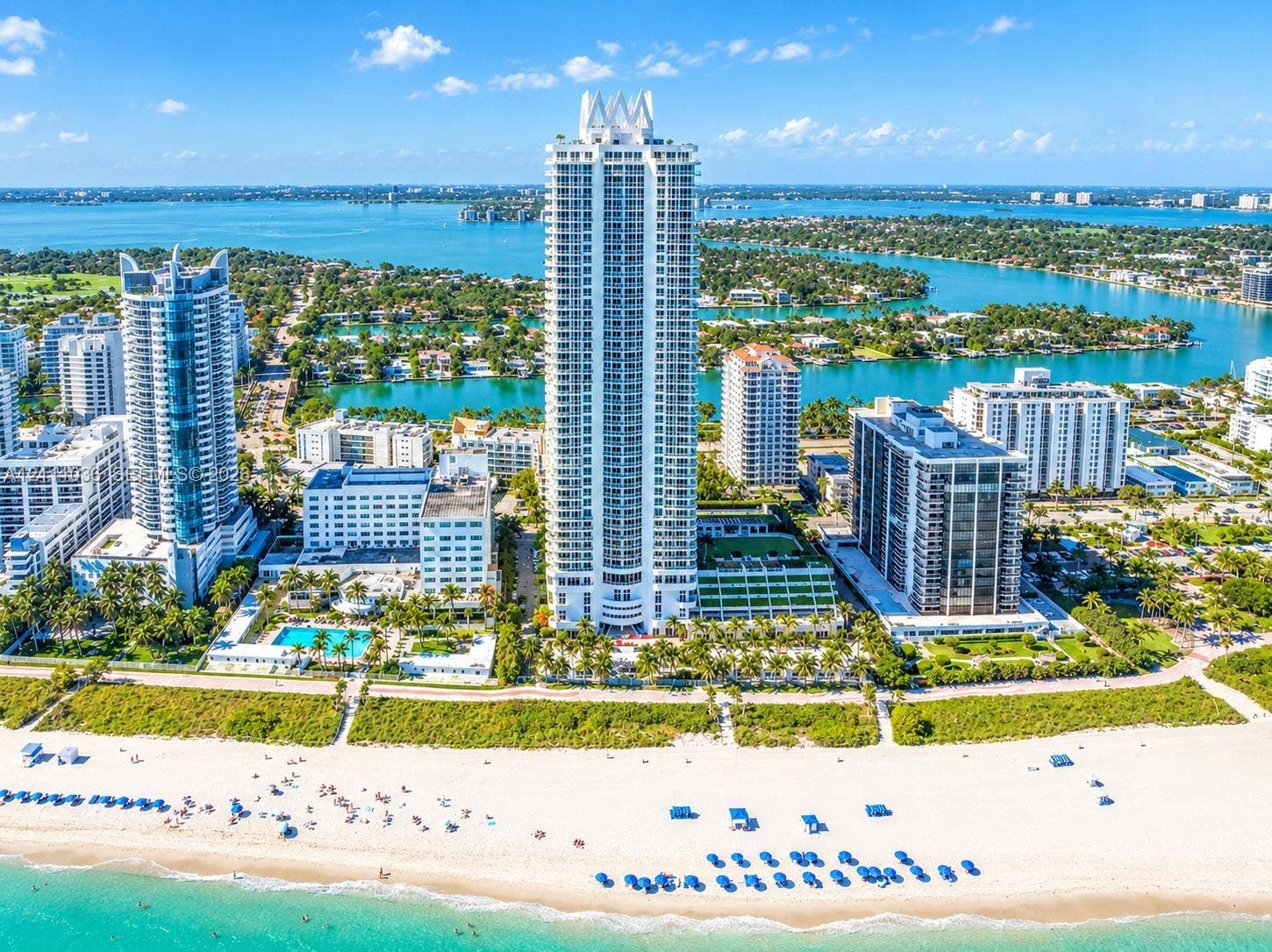 6365 Collins Avenue, #3811, Miami Beach, FL 33141 High-rise residential tower featuring a white concrete exterior, tiered architectural design, expansive glass balcony railings, direct sand beach access, and surrounding coastal waterfront scenery
