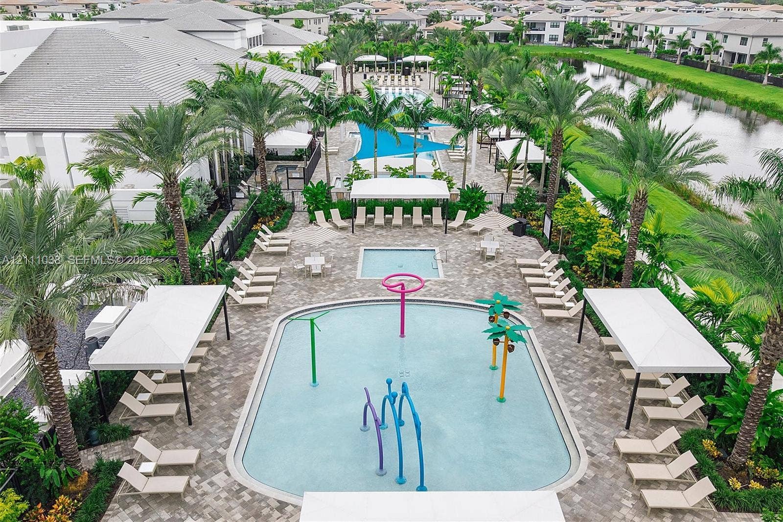 Resort-style aquatic area featuring a splash pad with water features, a separate spa, stone-paver decking, shaded cabanas, and mature palm trees