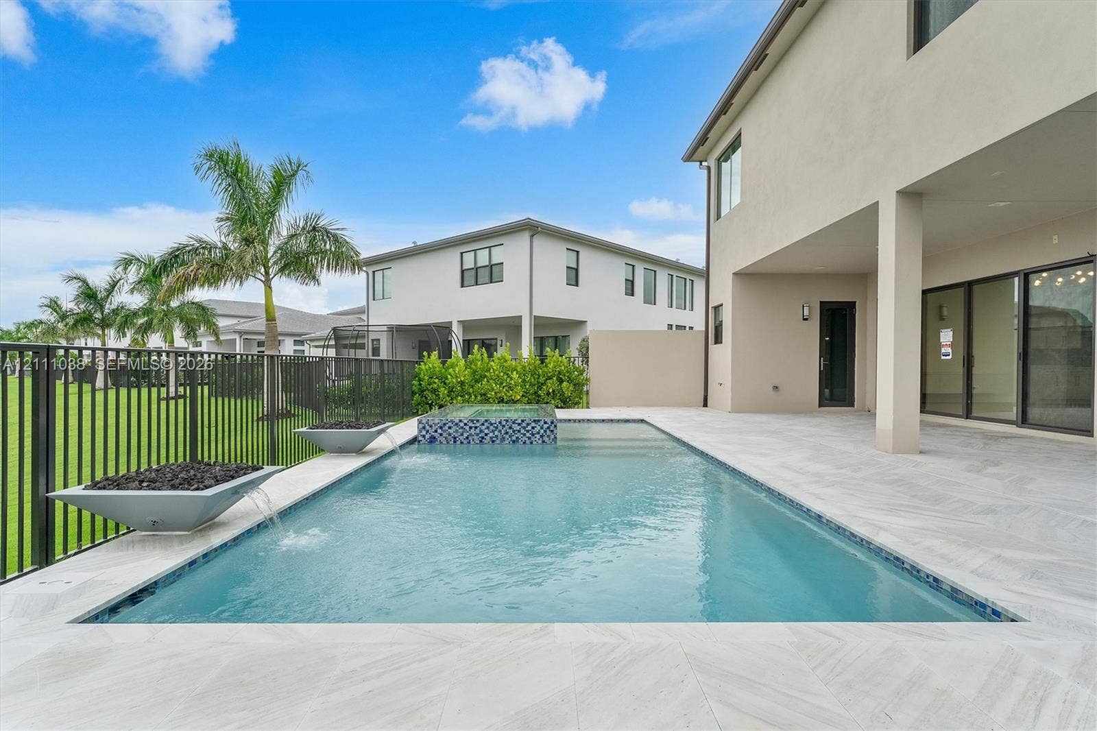 Swimming pool with a raised spa, decorative stone spillover bowls, neutral-toned tiled decking, and a black metal perimeter fence