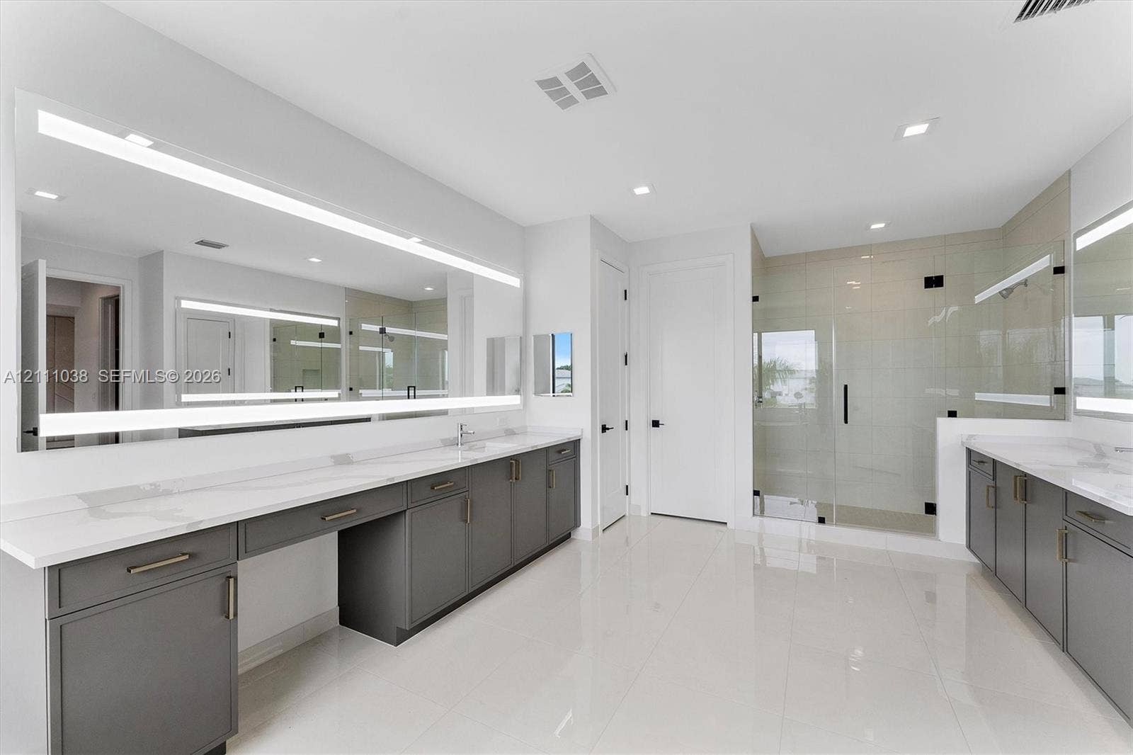 Dual vanities featuring charcoal cabinetry and white stone countertops, expansive wall-to-wall LED-backlit mirrors, polished tile flooring, and a glass-enclosed shower with tile surround