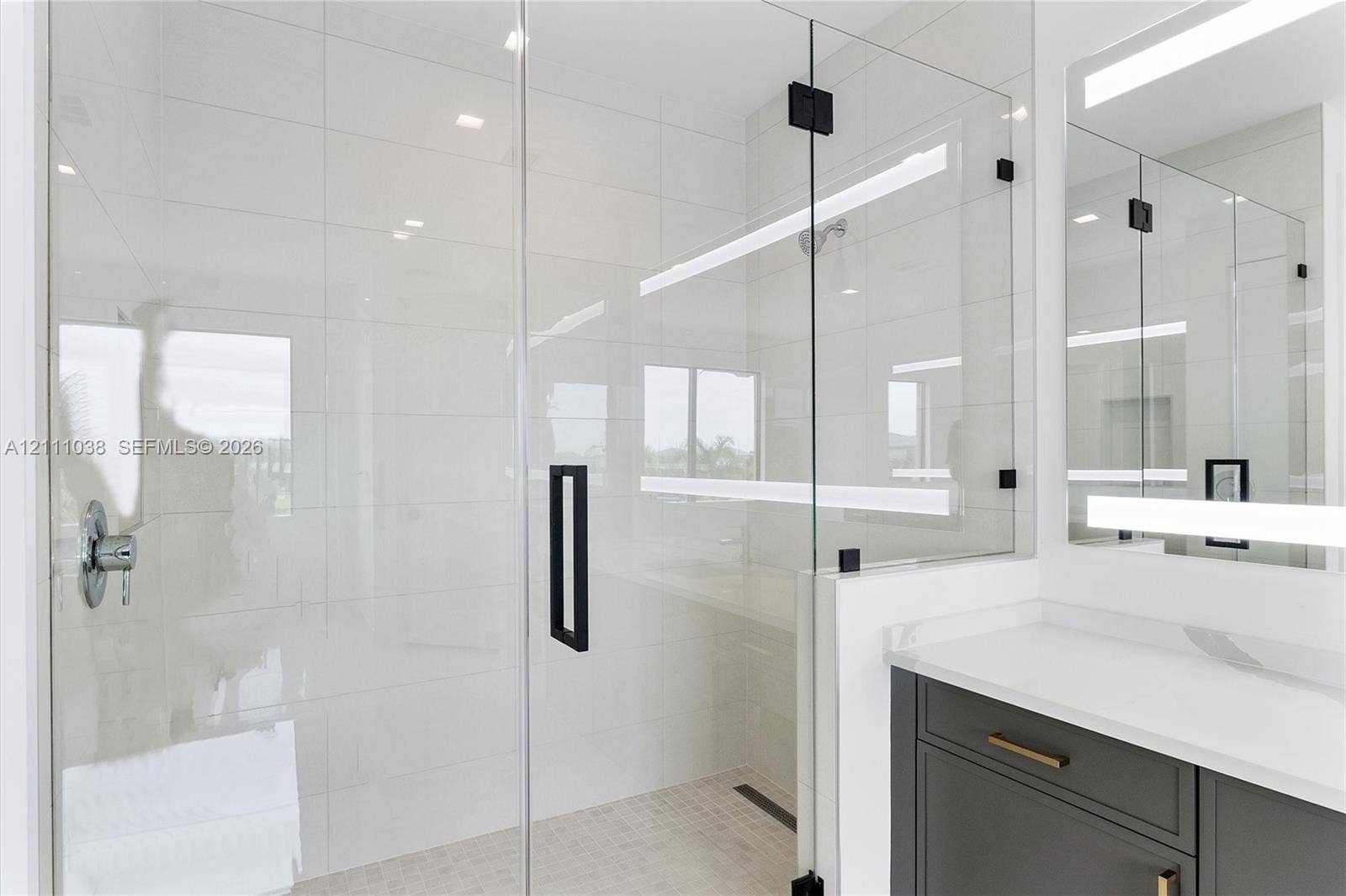 Walk-in shower with floor-to-ceiling tile, frameless glass enclosure, and matte black hardware, featuring a modern vanity with a white countertop and integrated linear LED mirror lighting