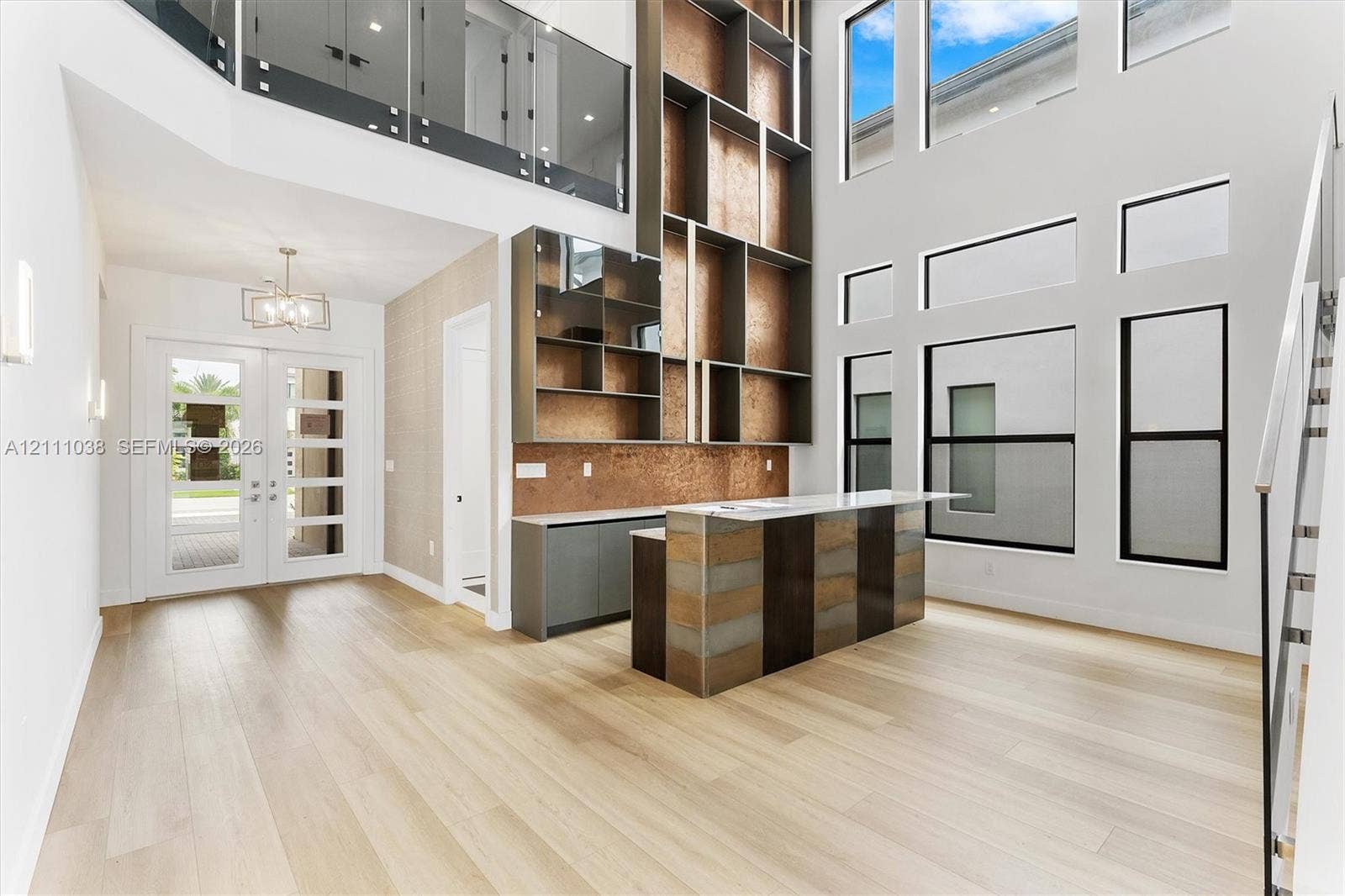 Double-height ceiling, floor-to-ceiling windows, custom built-in cabinetry with integrated shelving, stone-topped island, and light-toned wood-finish flooring