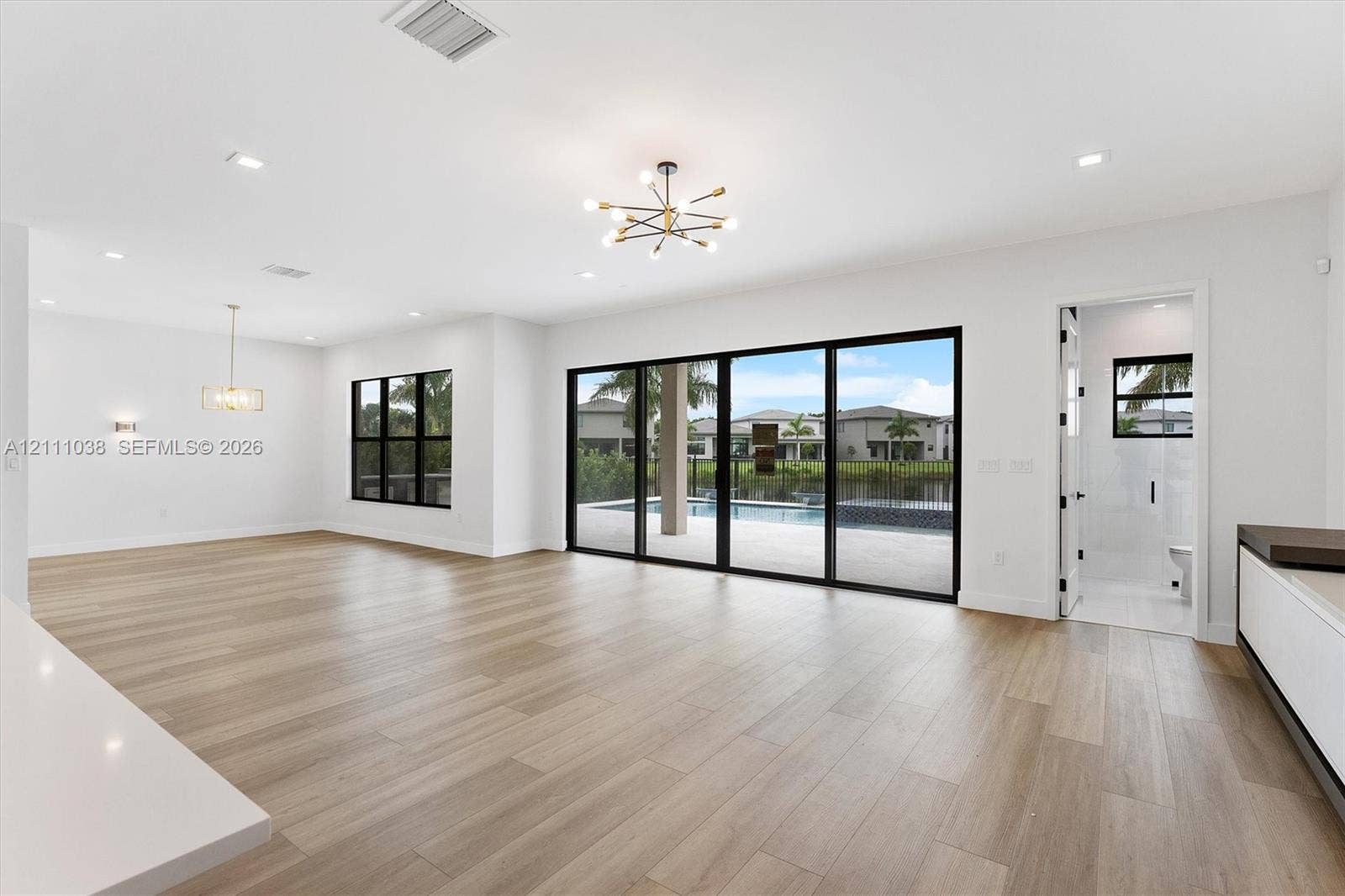 Open-concept interior featuring light wood-finish flooring, multi-panel sliding glass doors with black frames, recessed lighting, and modern multi-arm chandeliers