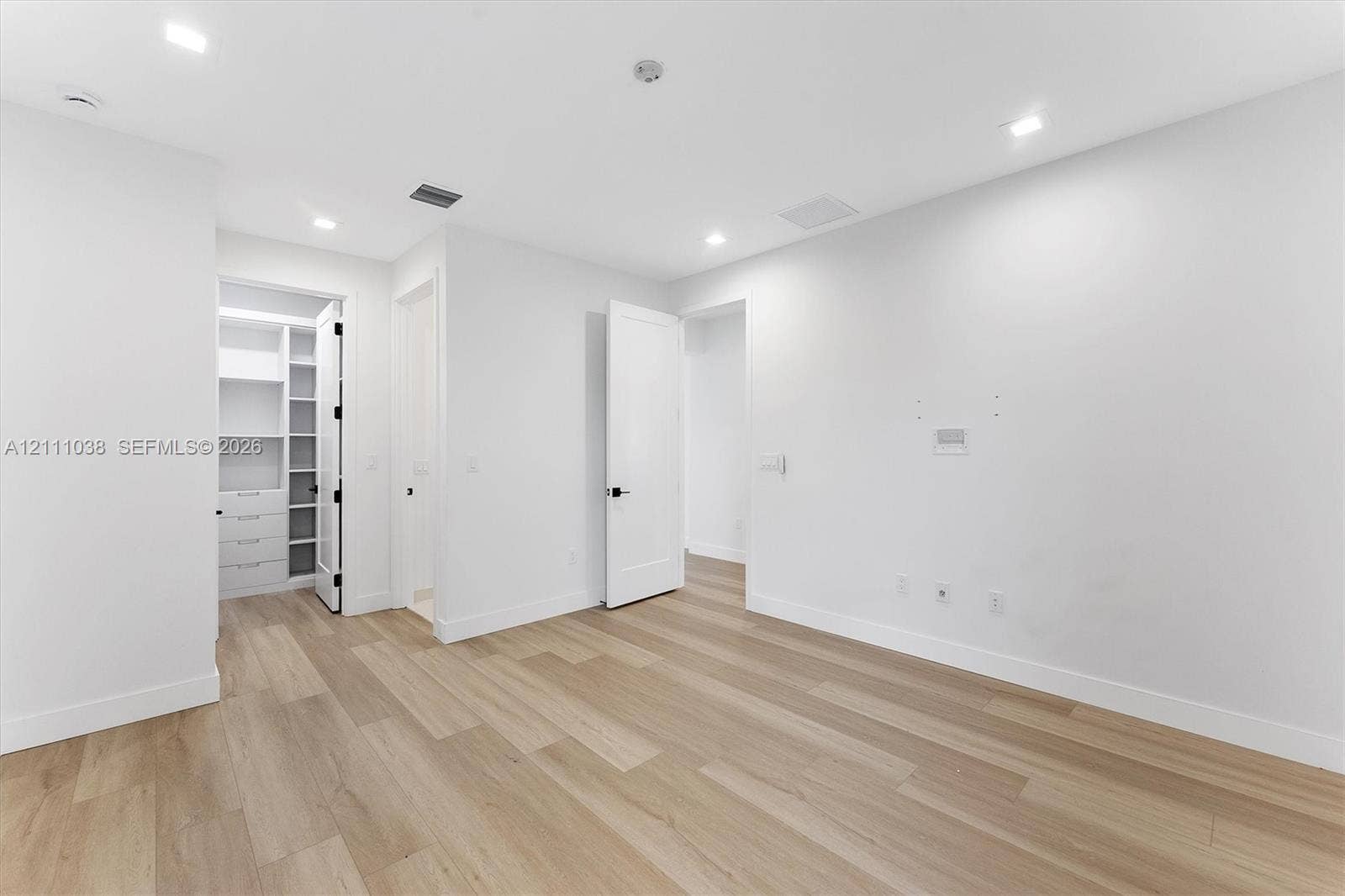 Neutral-toned space featuring wood-finish flooring, recessed lighting, shaker-style doors with matte black hardware, and an integrated walk-in closet with built-in shelving