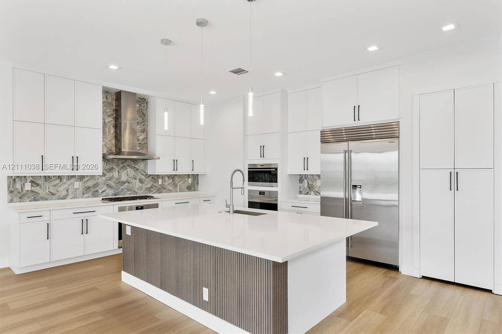 Kitchen featuring crisp white cabinetry, a center island with a ribbed wood-finish base, quartz countertops, a patterned stone tile backsplash, and stainless steel appliances
