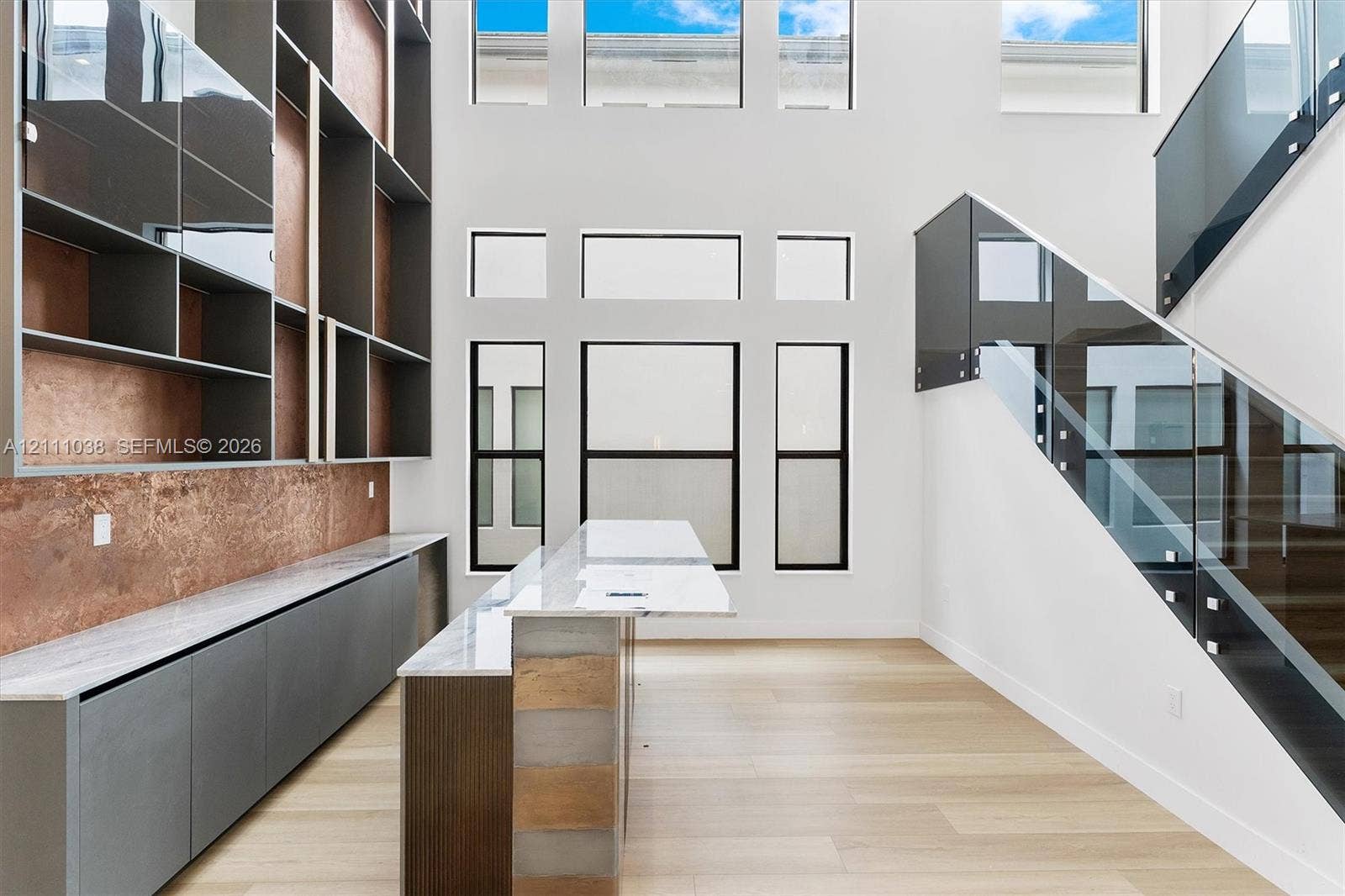 Double-height ceiling with multi-level windows, floor-to-ceiling built-in shelving with integrated stone-finish backsplash, wood-finish flooring, and a glass-panel staircase railing