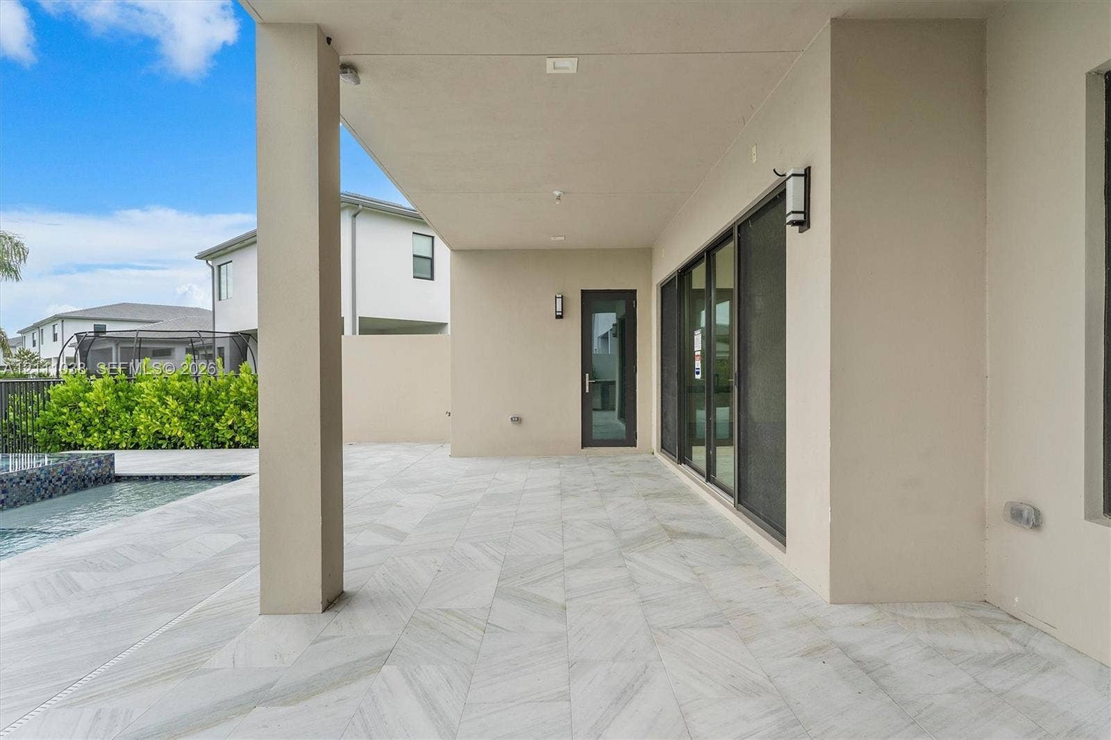 Covered outdoor patio featuring large-format tile flooring, recessed ceiling lighting, wall-mounted fixtures, and a glass-paneled entry door, situated beside a pool with tile surround