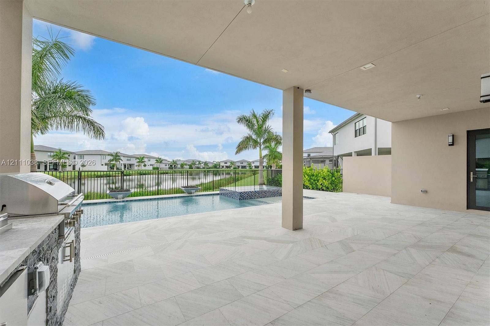 Covered lanai featuring large-format tile flooring, outdoor kitchen with stainless steel grill, in-ground pool with spillover spa, and metal fencing
