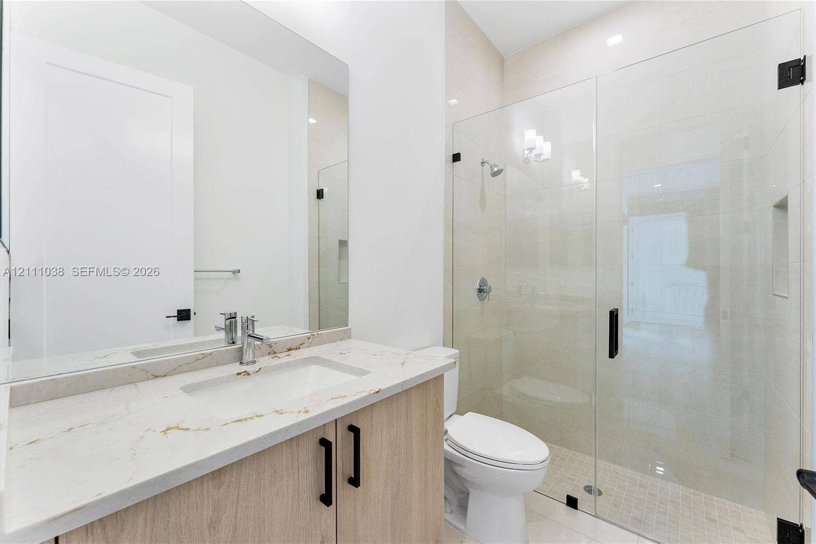 Bathroom featuring a vanity with light wood-finish cabinetry, a stone-surface countertop with an integrated rectangular sink, a large wall-mounted mirror, and a glass-enclosed shower with a recessed wall niche