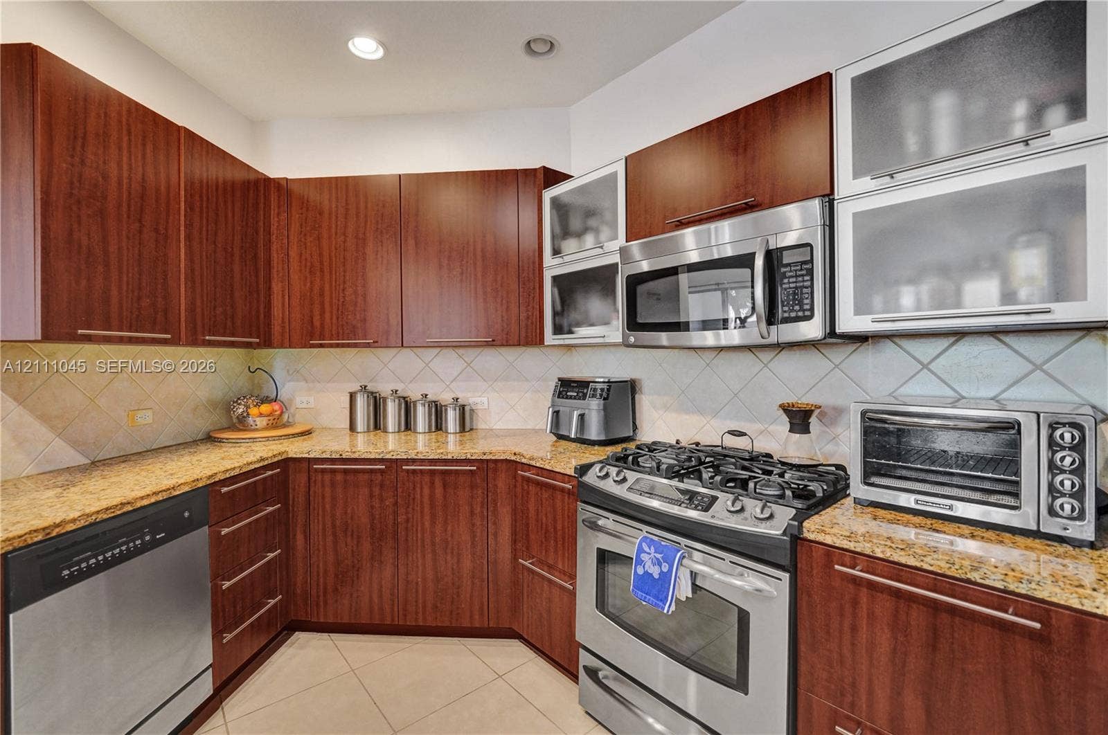 2901 NW 126th Avenue, #2-225, Sunrise, FL 33323 Kitchen featuring warm-tone cabinetry, natural stone countertops, diagonal tile backsplash, stainless steel appliances, and glass-front upper cabinets