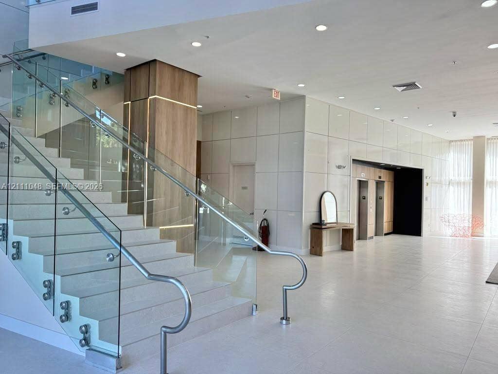 2775 NE 187th Street, #408, Aventura, Aventura, FL 33180 Lobby area featuring a wide staircase with glass railings and metal handrails, a central pillar with integrated accent lighting and wood-finish paneling, large-format tiled flooring, and high-gloss wall panels