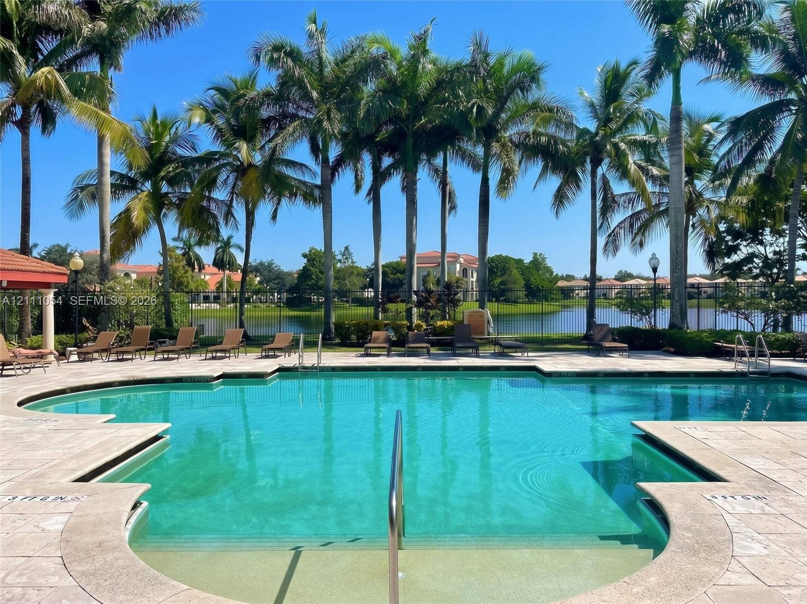 4180 San Marino Boulevard, #106, West Palm Beach, FL 33409 Swimming pool with stone paver decking, metal perimeter fencing, and surrounding mature palm trees overlooking a lake