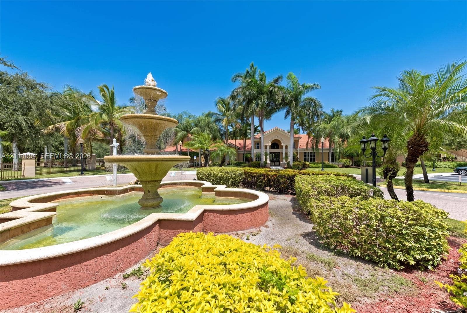 4180 San Marino Boulevard, #106, West Palm Beach, FL 33409 Tiered stone water fountain featuring a decorative basin, surrounded by professional landscaping, manicured hedges, and mature palm trees