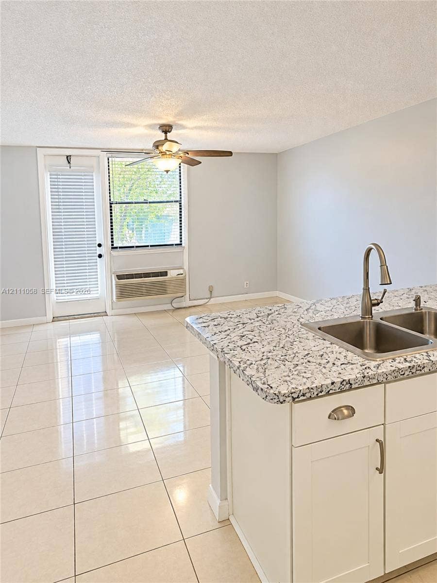 Kitchen island featuring a stone-look countertop, undermount double-basin stainless steel sink, polished chrome faucet, white Shaker-style cabinetry, and light-toned tile flooring