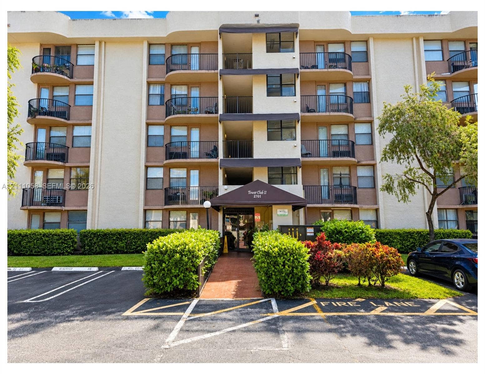 Multi-story residential building exterior featuring curved private balconies, metal railings, and a covered entryway with a brick-paved walkway