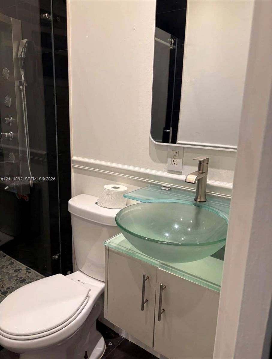 17631 32nd Court, Miami Gardens, FL 33056 Frosted glass vessel sink with a glass countertop, chrome-finish faucet, white cabinetry with metal hardware, and a glass-enclosed shower featuring a multi-head fixture