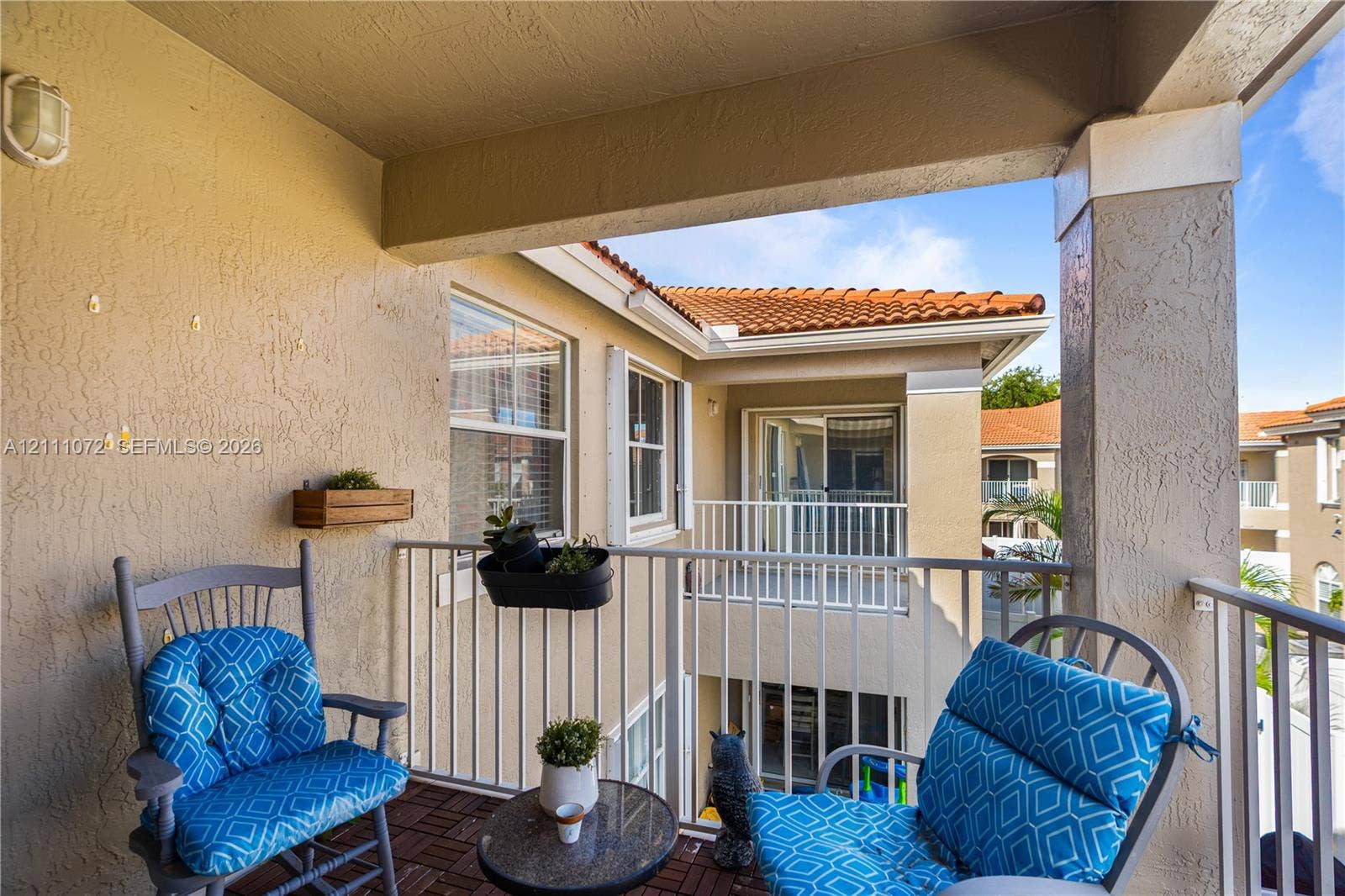 Covered balcony featuring textured stucco walls, tiled flooring, metal railings, and a wall-mounted light fixture