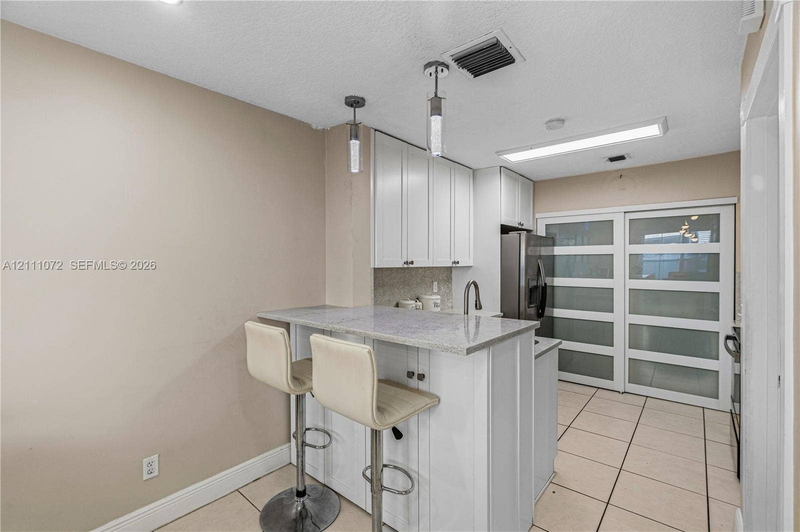 Kitchen featuring white shaker-style cabinetry, stone-surface breakfast bar with integrated sink, stainless steel refrigerator, and frosted-glass panel sliding doors, finished with tile flooring and dual pendant lighting