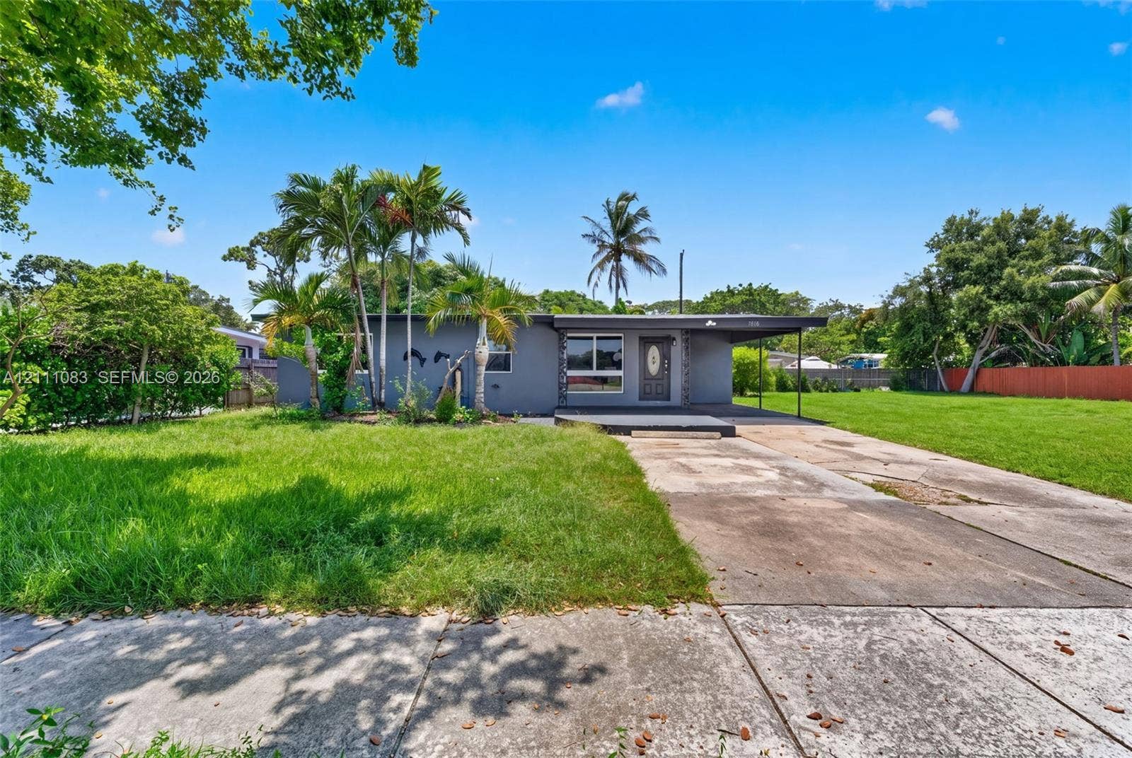 Single-story exterior featuring a flat roofline, attached covered carport, concrete driveway, and established palm trees
