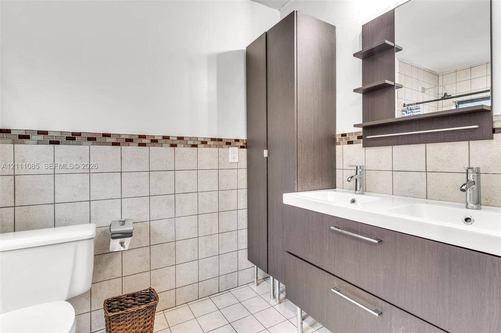 Double vanity with wood-finish cabinetry, integrated storage tower, dual metal faucets, and wall-mounted mirror with shelving, complemented by tiled wall accents