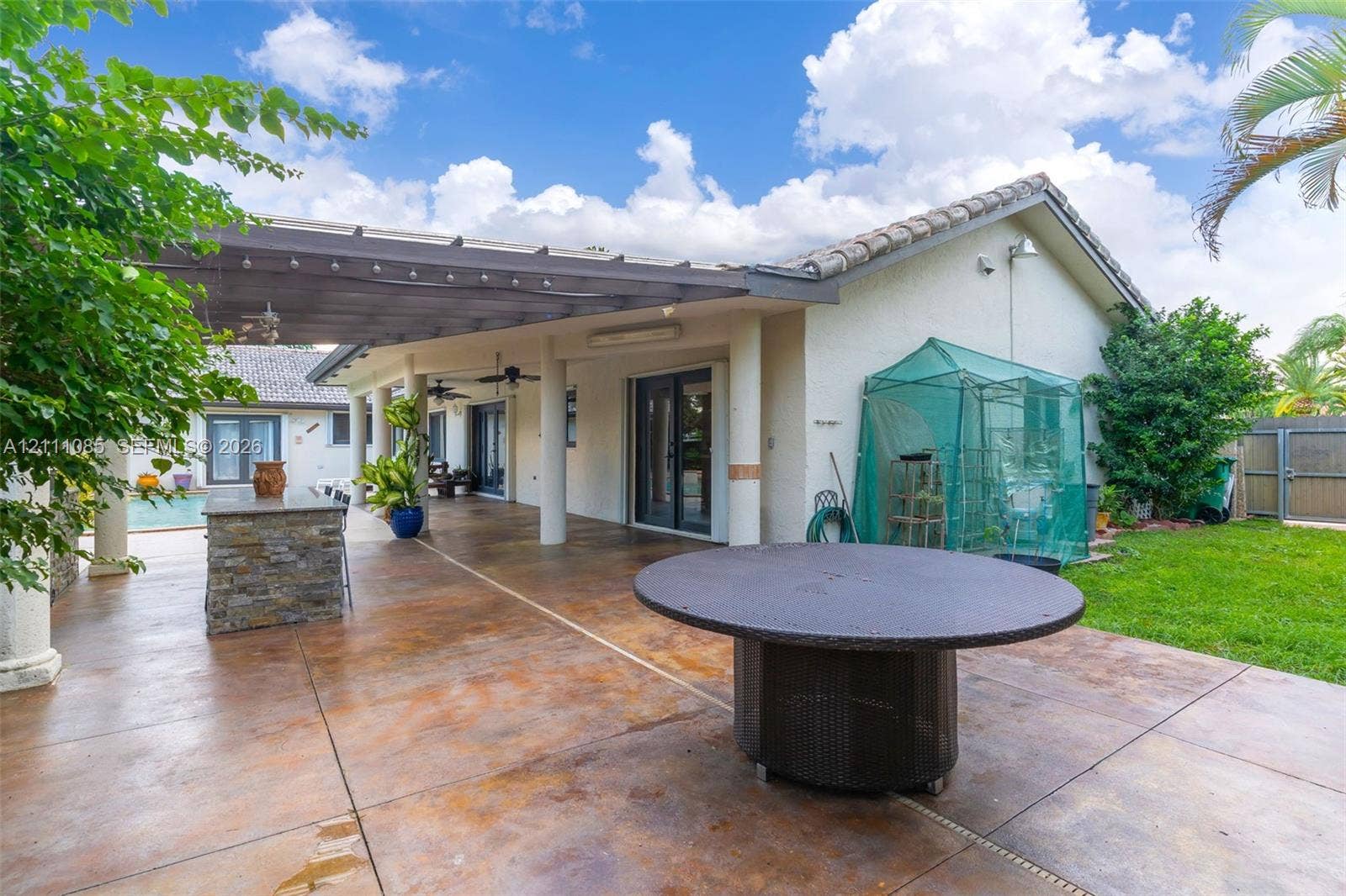 Covered concrete patio featuring a stone-clad outdoor kitchen, structural columns, ceiling fans, and built-in lighting